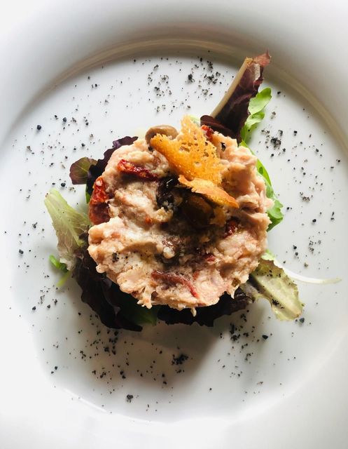 Antipasto di insalata di tonno su un letto di lattuga, guarnito con pomodori secchi, olive e un cracker su un piatto bianco.