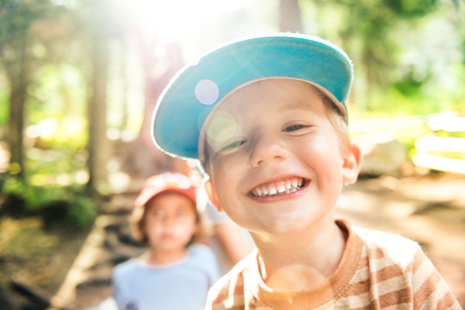 Smiling young boy in blue cap, sunlight, with another child in the background on a path in a forest.