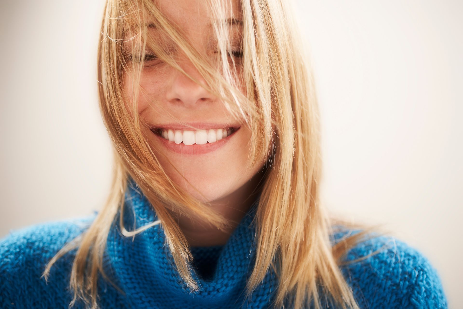 Blonde woman smiling, strands of hair in her face, wearing a blue sweater.