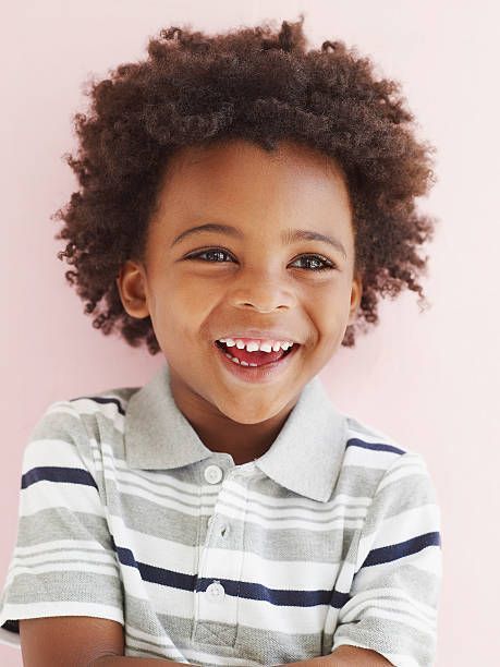 Smiling Black boy with curly hair, wearing a striped shirt, stands against a pink background.