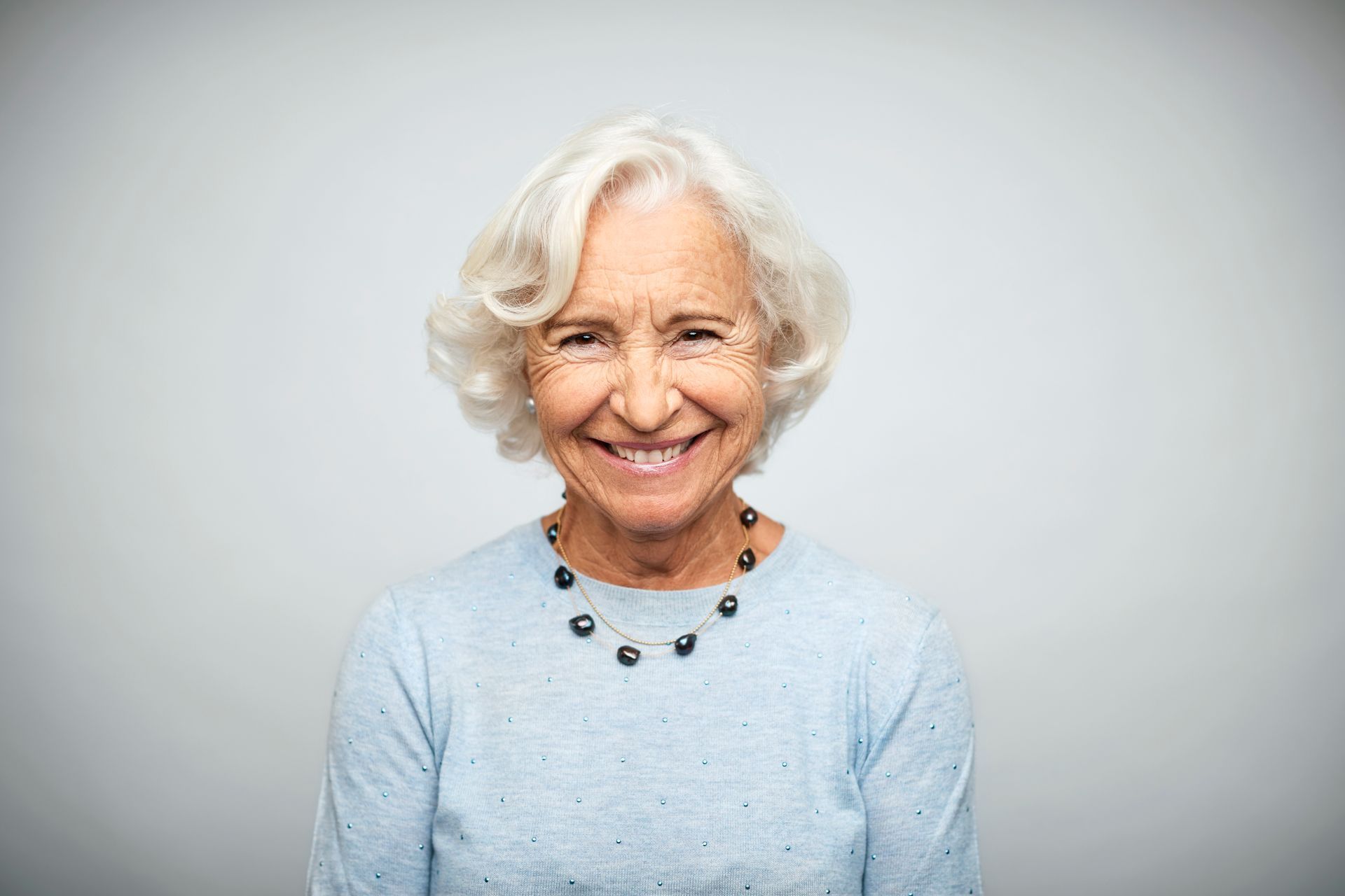 Smiling older woman with white curly hair, wearing a blue sweater and beaded necklace.