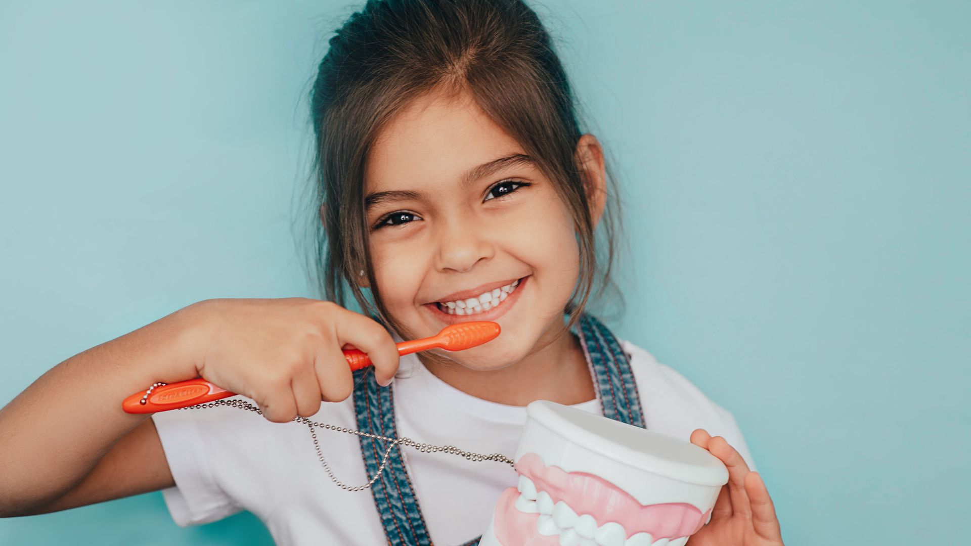 Young girl with a bright smile, holding an orange toothbrush and teeth model, against a blue background.