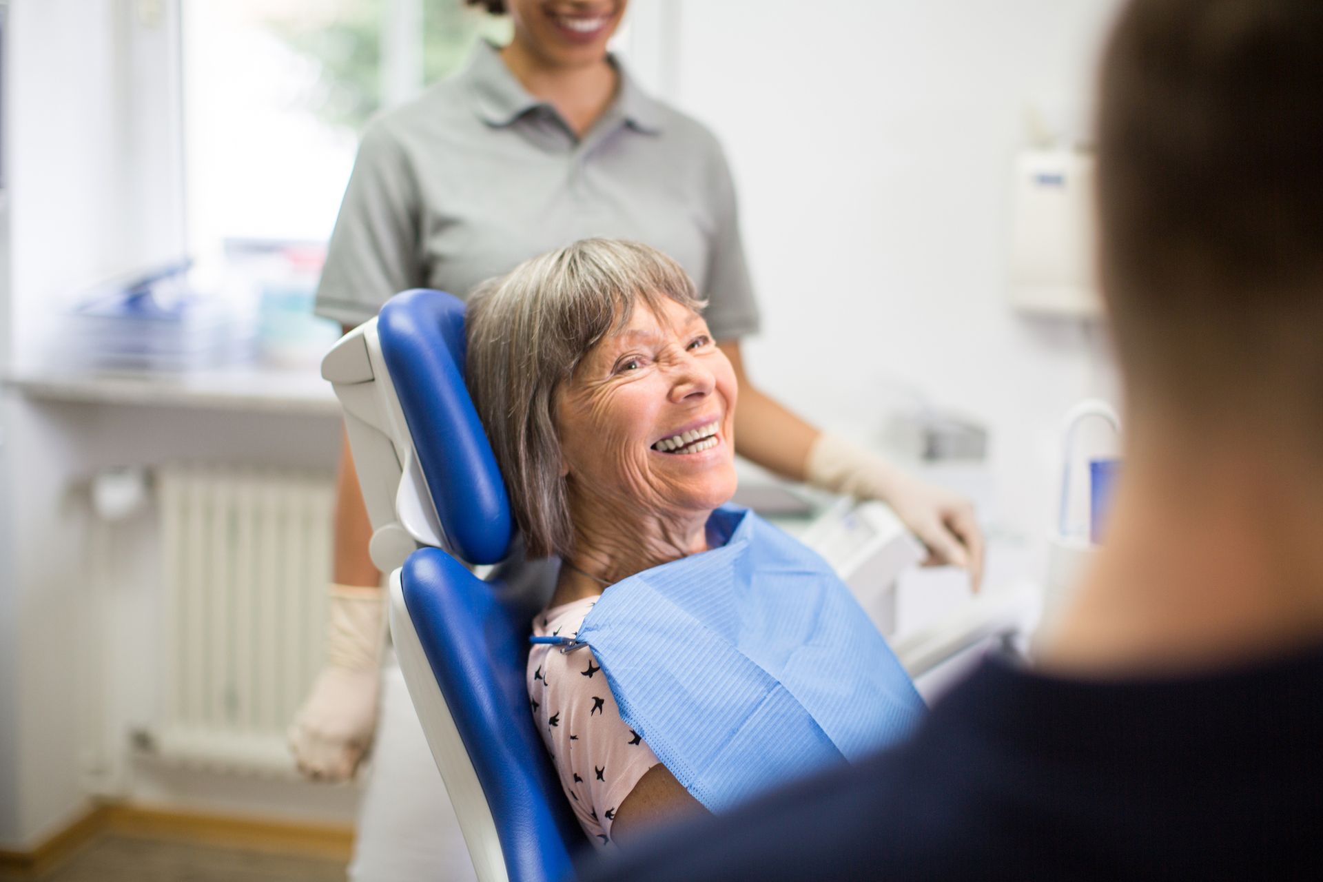 Smiling elderly woman in dentist chair, dental assistant and dentist attending.