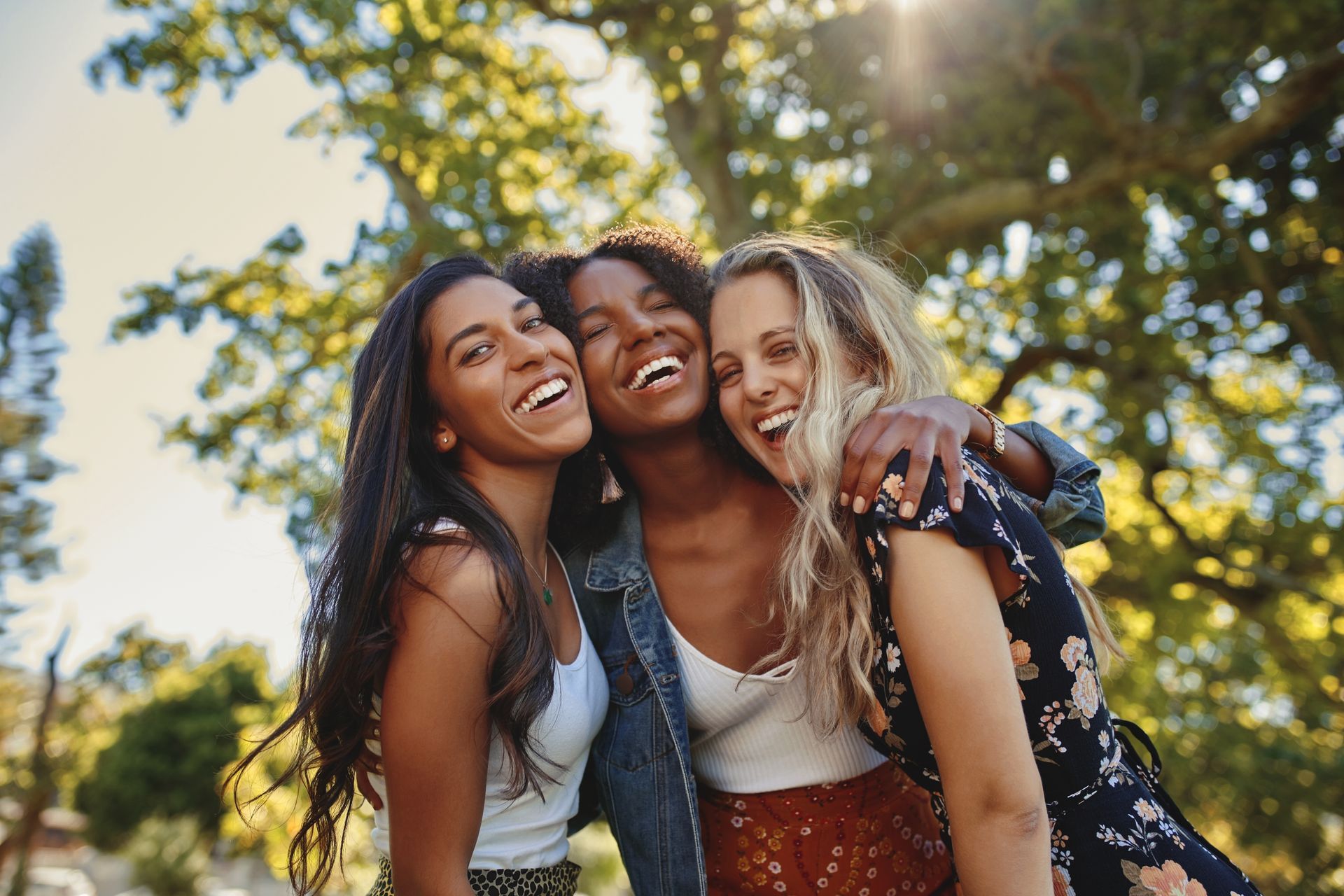 Three diverse women smiling and embracing outdoors in sunlight.