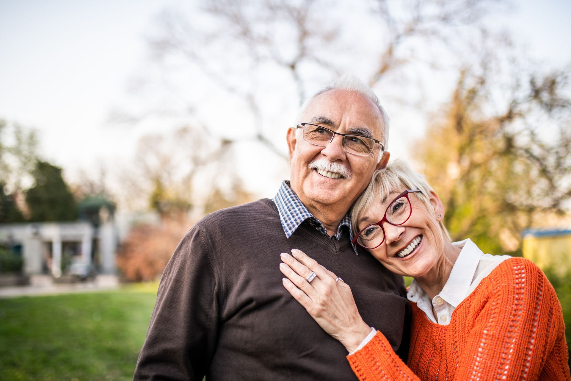 Smiling elderly couple outdoors, woman leaning on man. Green grass, trees.