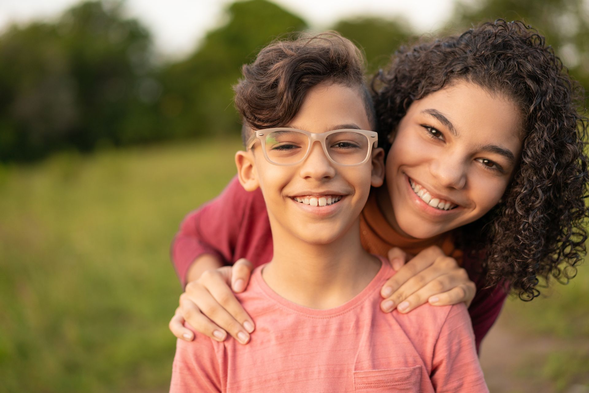 Two smiling youth, an older girl with curly hair hugs a boy with glasses and a modern haircut outdoors.