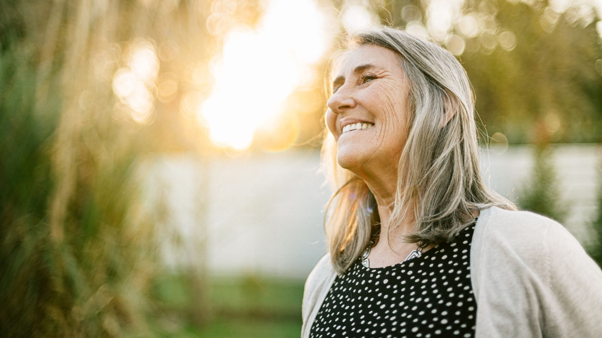 Smiling elderly woman with gray hair looking up, outdoors in the sunlight.