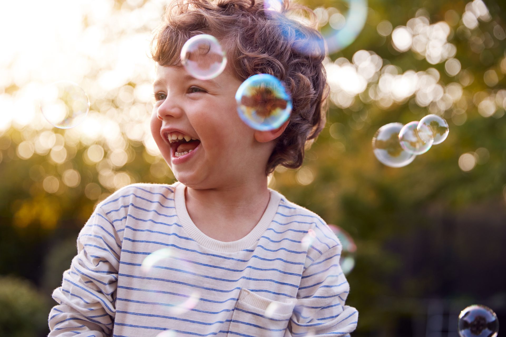 Happy child with curly hair, laughing as soap bubbles float around them outdoors.