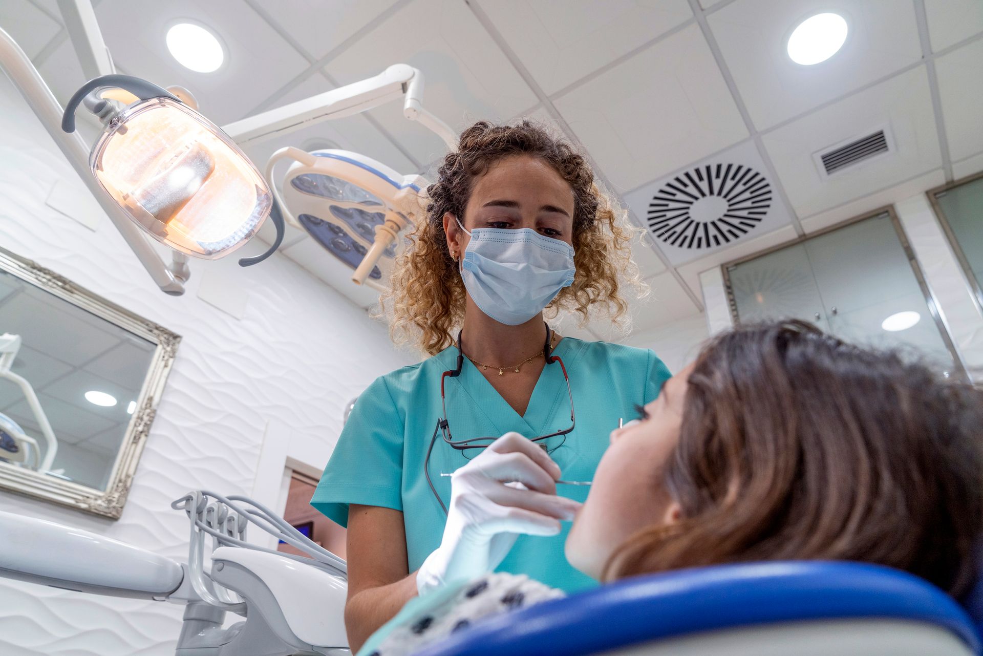 Dentist in blue scrubs examines patient's teeth. Dentist wearing mask and gloves, in dental office.