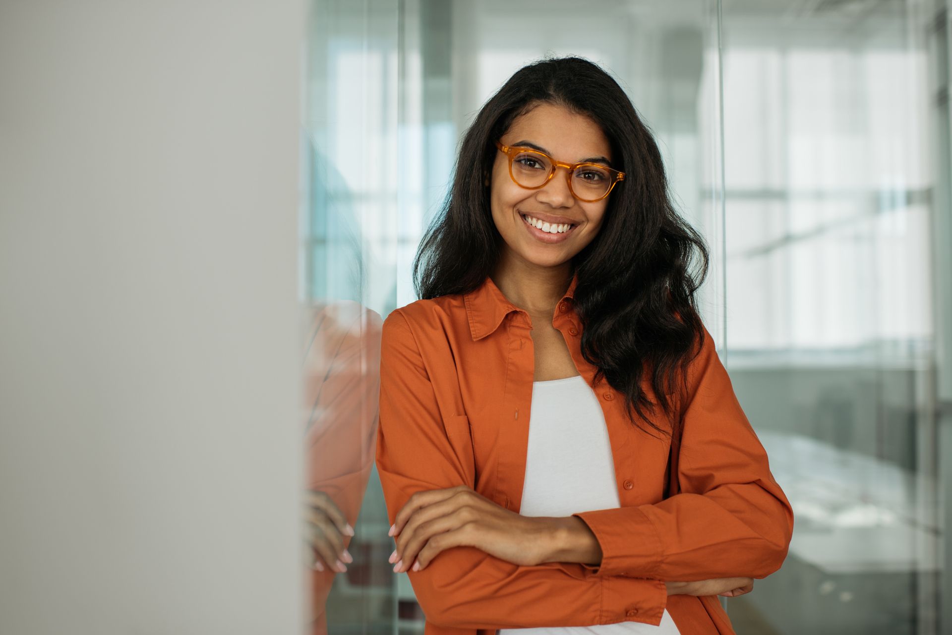 Smiling woman with crossed arms, wearing orange shirt and glasses, leaning against a wall.