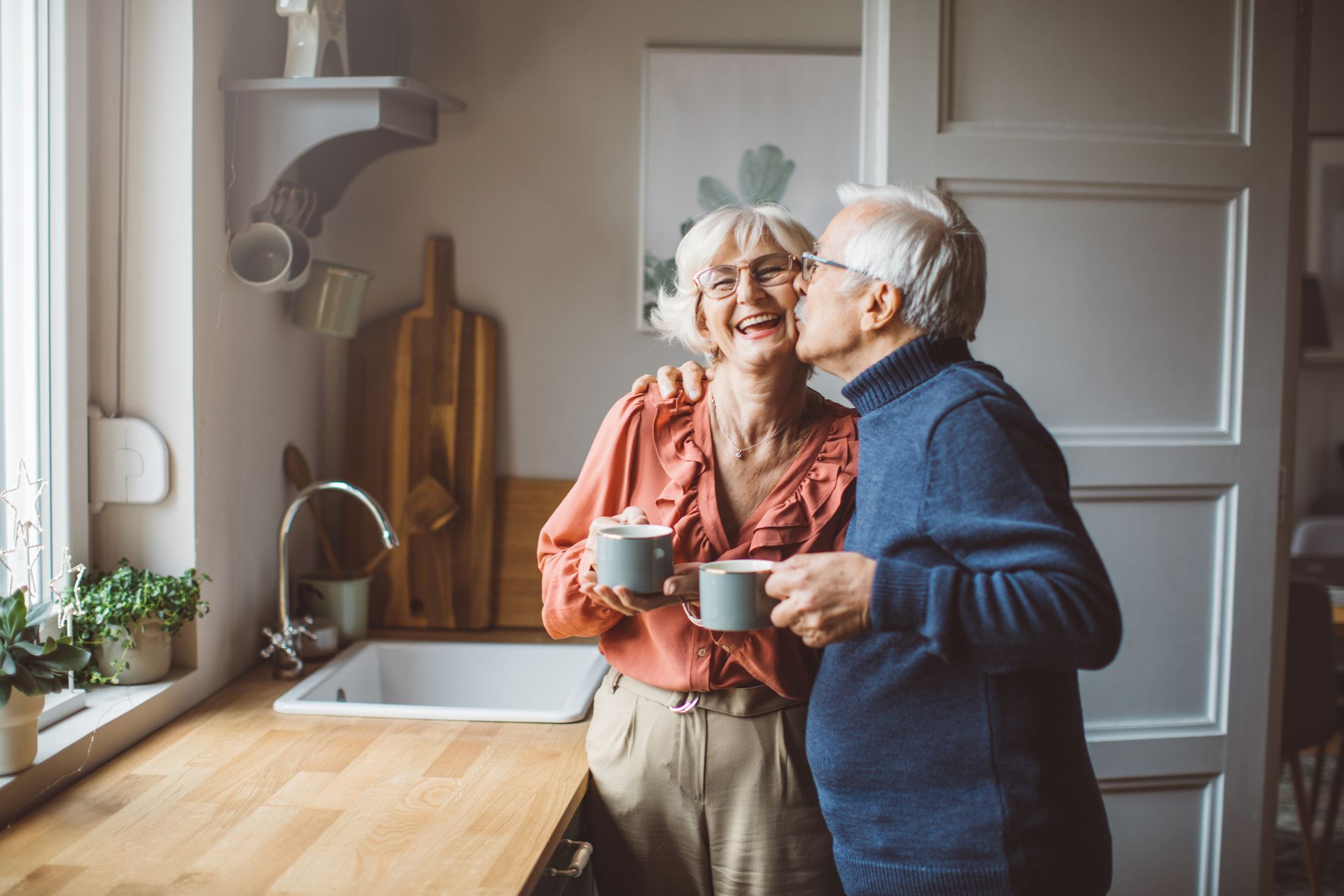 Older couple in kitchen, man kissing woman's cheek while holding mugs. Happy expressions.