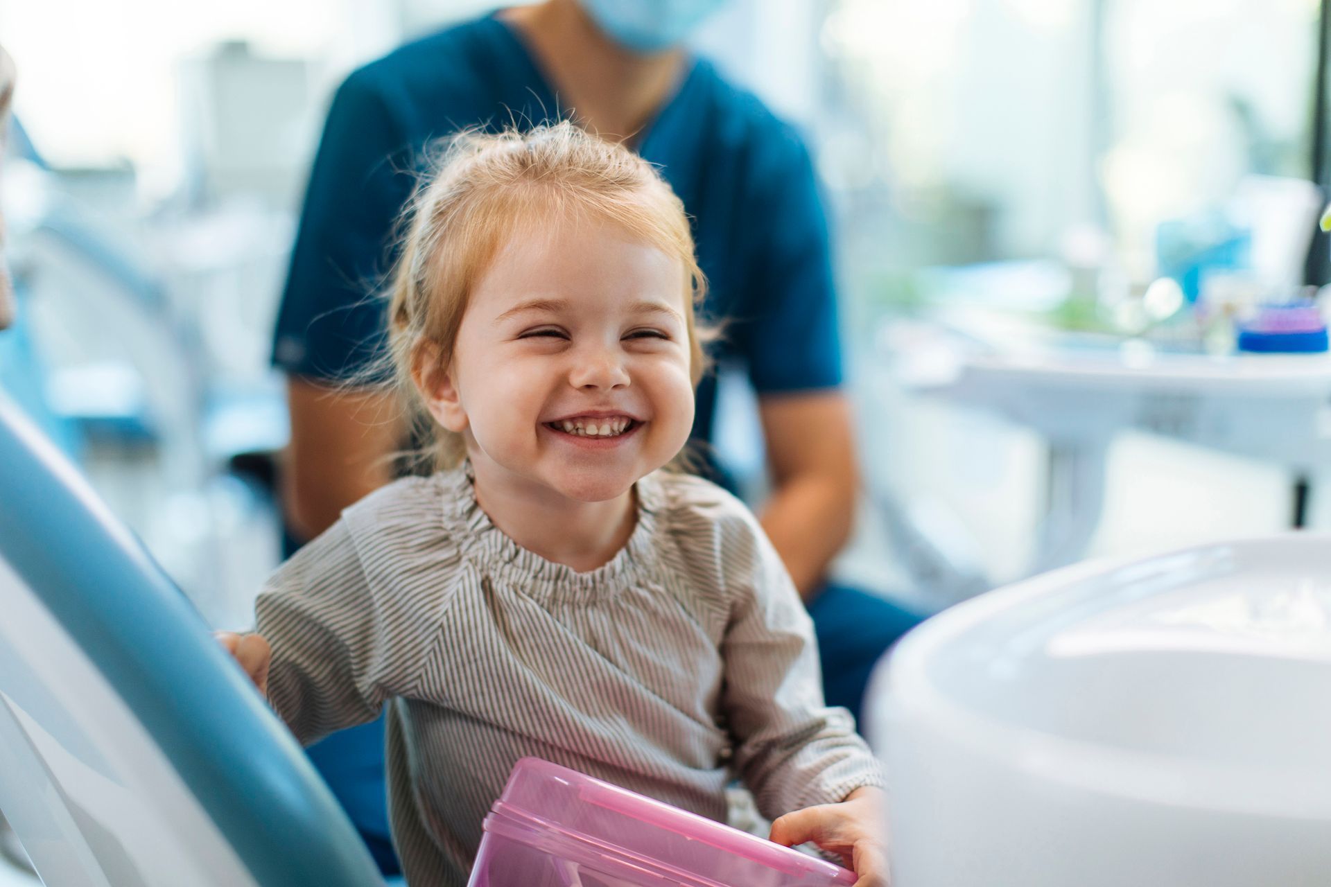 Smiling toddler at dentist, holding a pink container, blue chair, dentist in background.