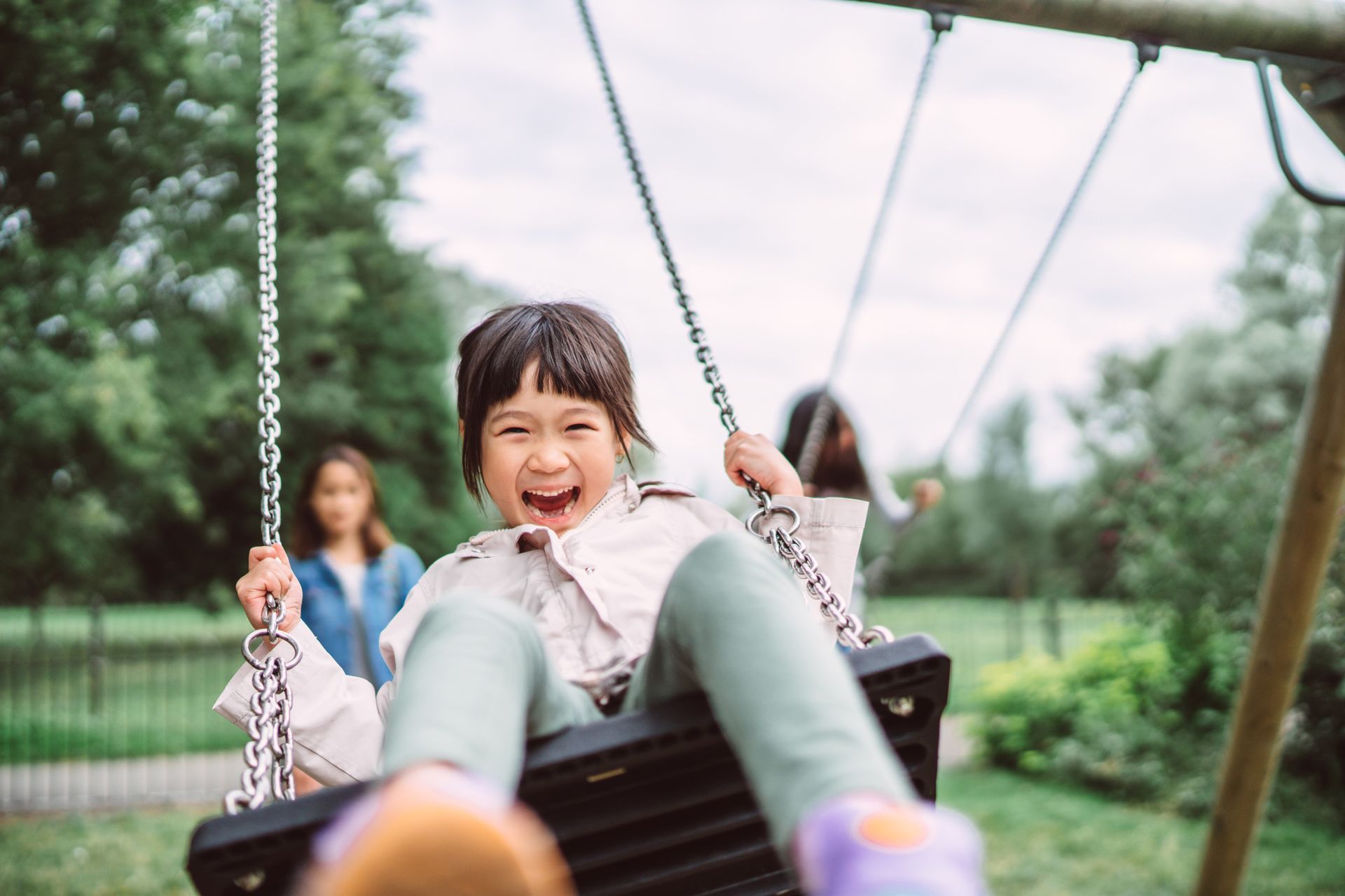 Girl with open mouth laughing on swing; another person pushing in background.