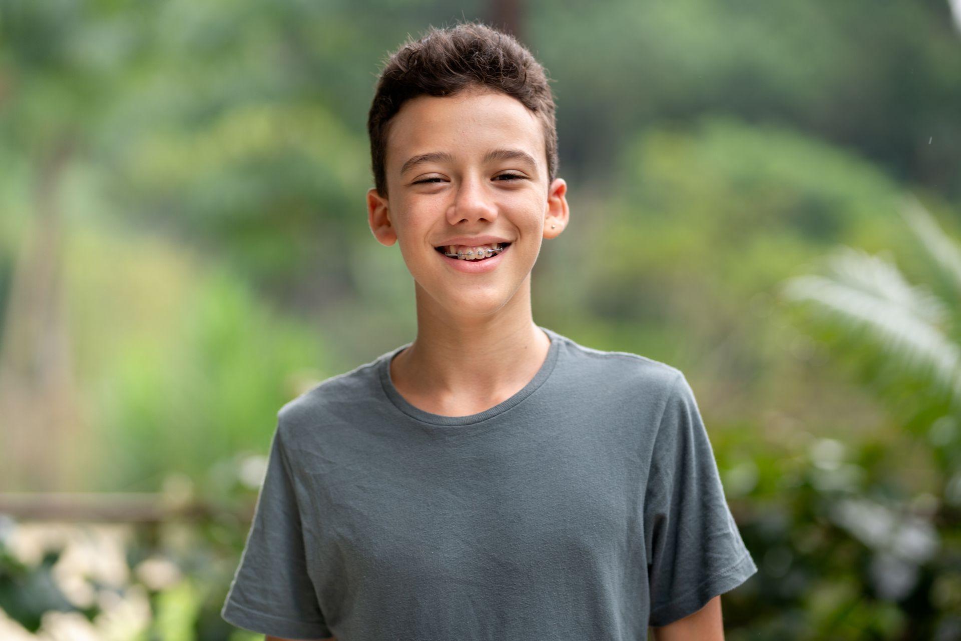 Boy with braces smiles in front of a blurred green background, wearing a gray t-shirt.