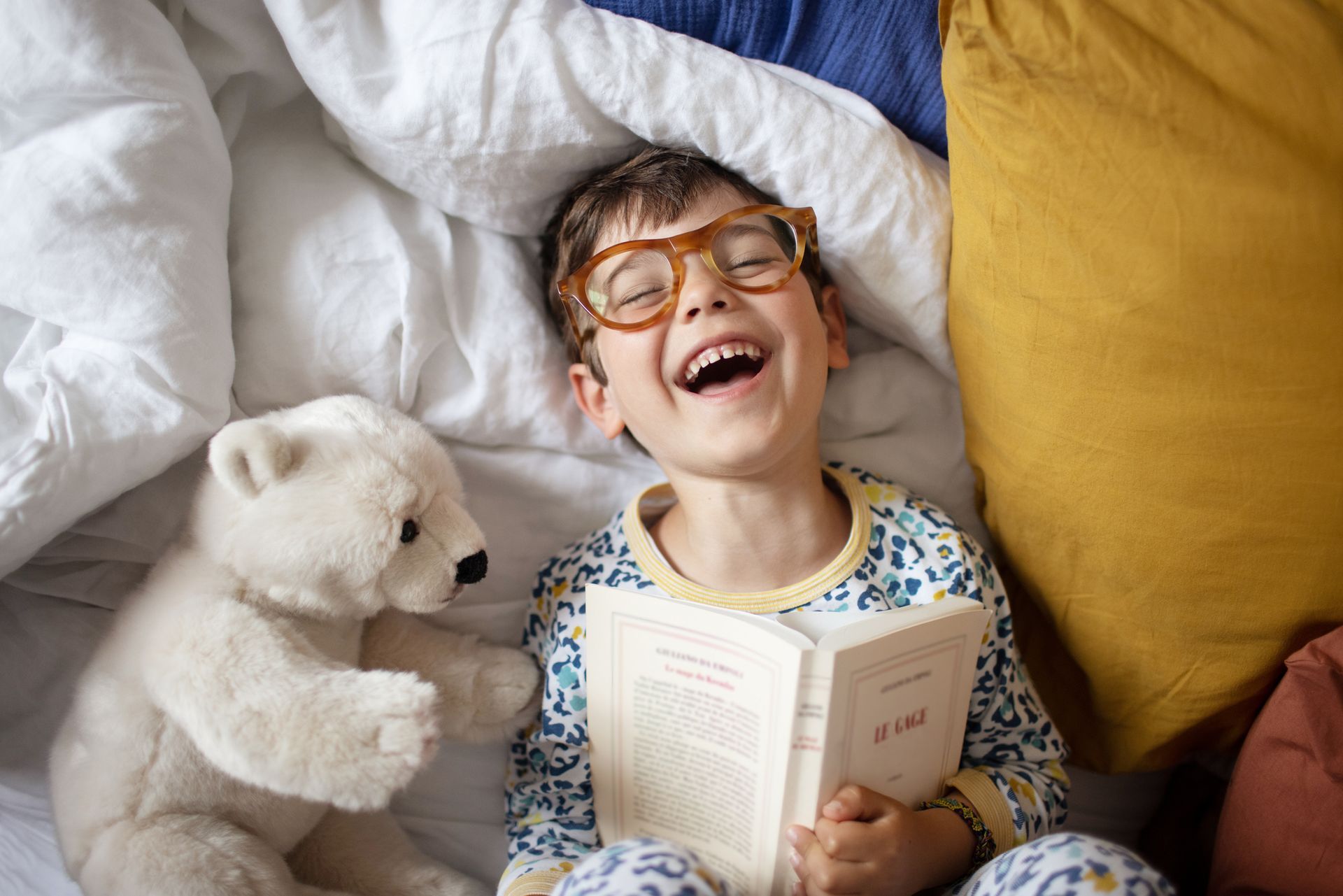 Boy wearing glasses, laughing while reading in bed with a teddy bear and book.