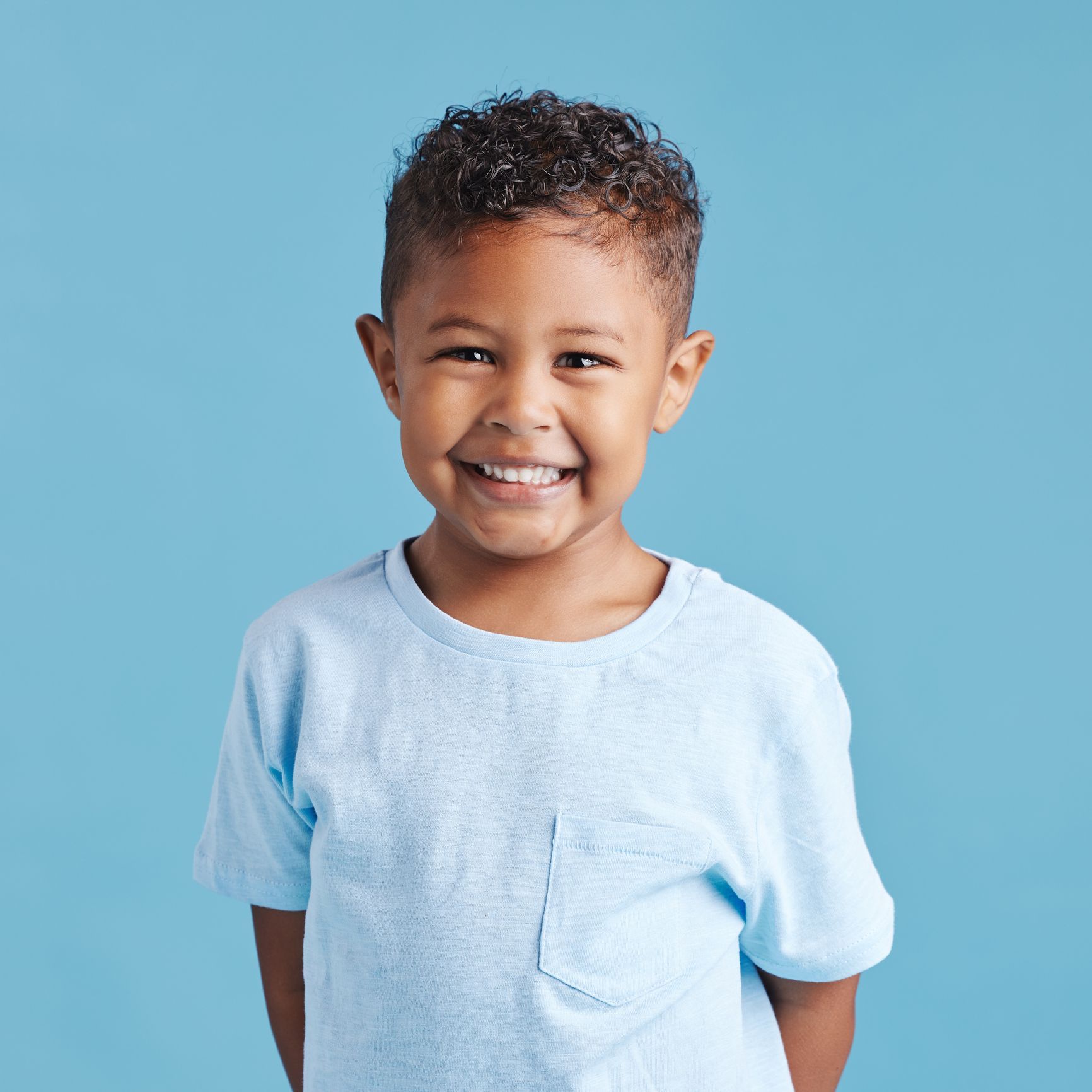 Smiling young boy with curly hair wearing a blue t-shirt against a blue background.
