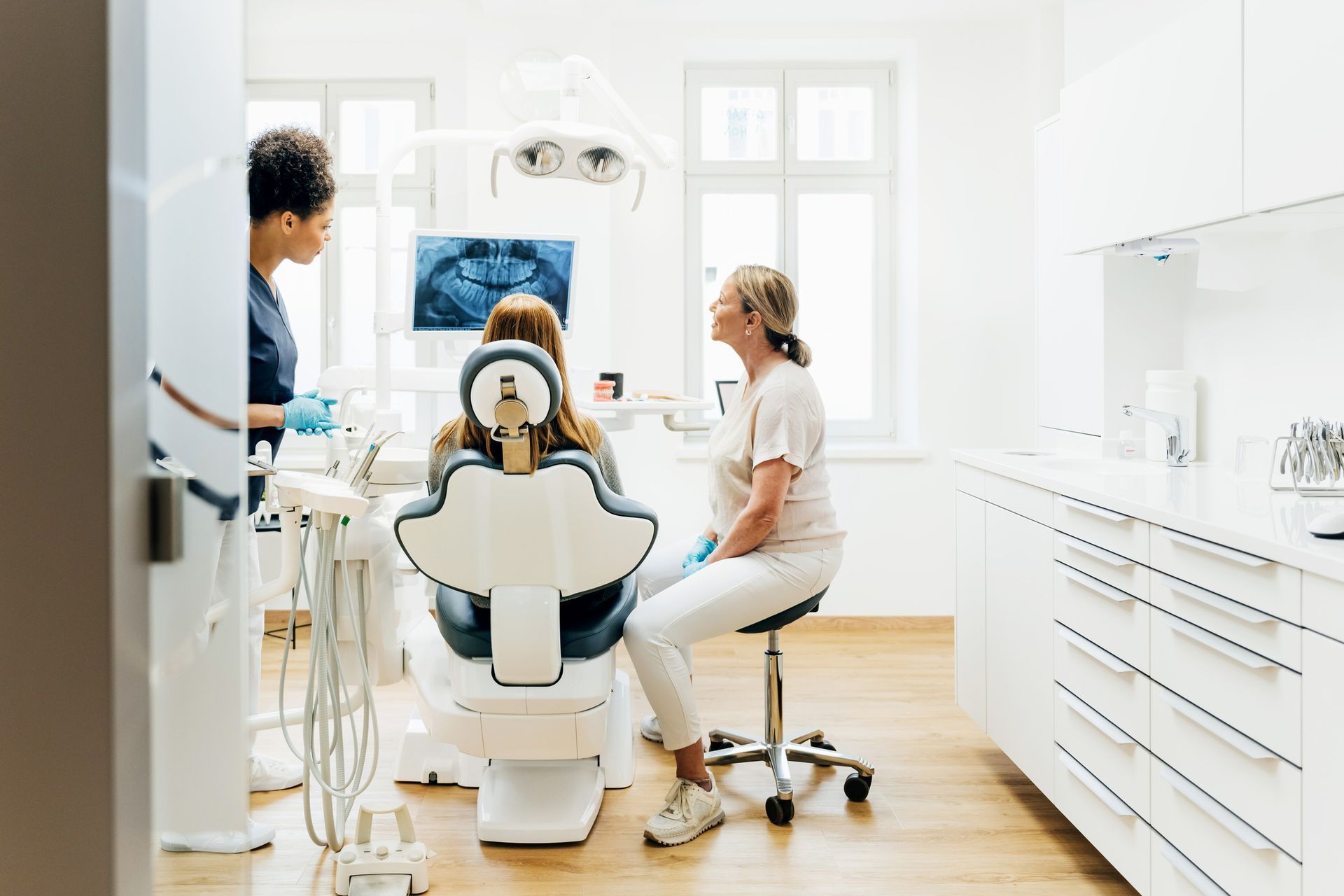 Dentist examining patient's x-ray in dental office, assistant present. Brightly lit room, modern equipment.