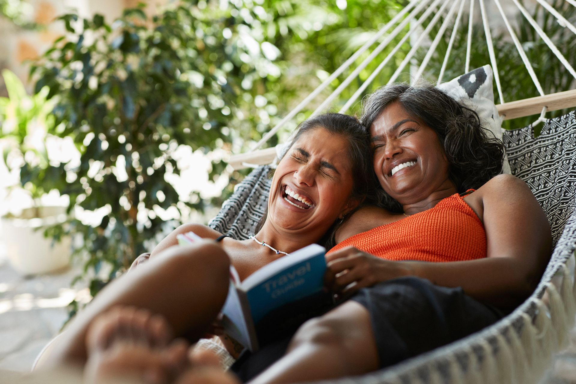 Two women laughing in a hammock, one reading a book outdoors.