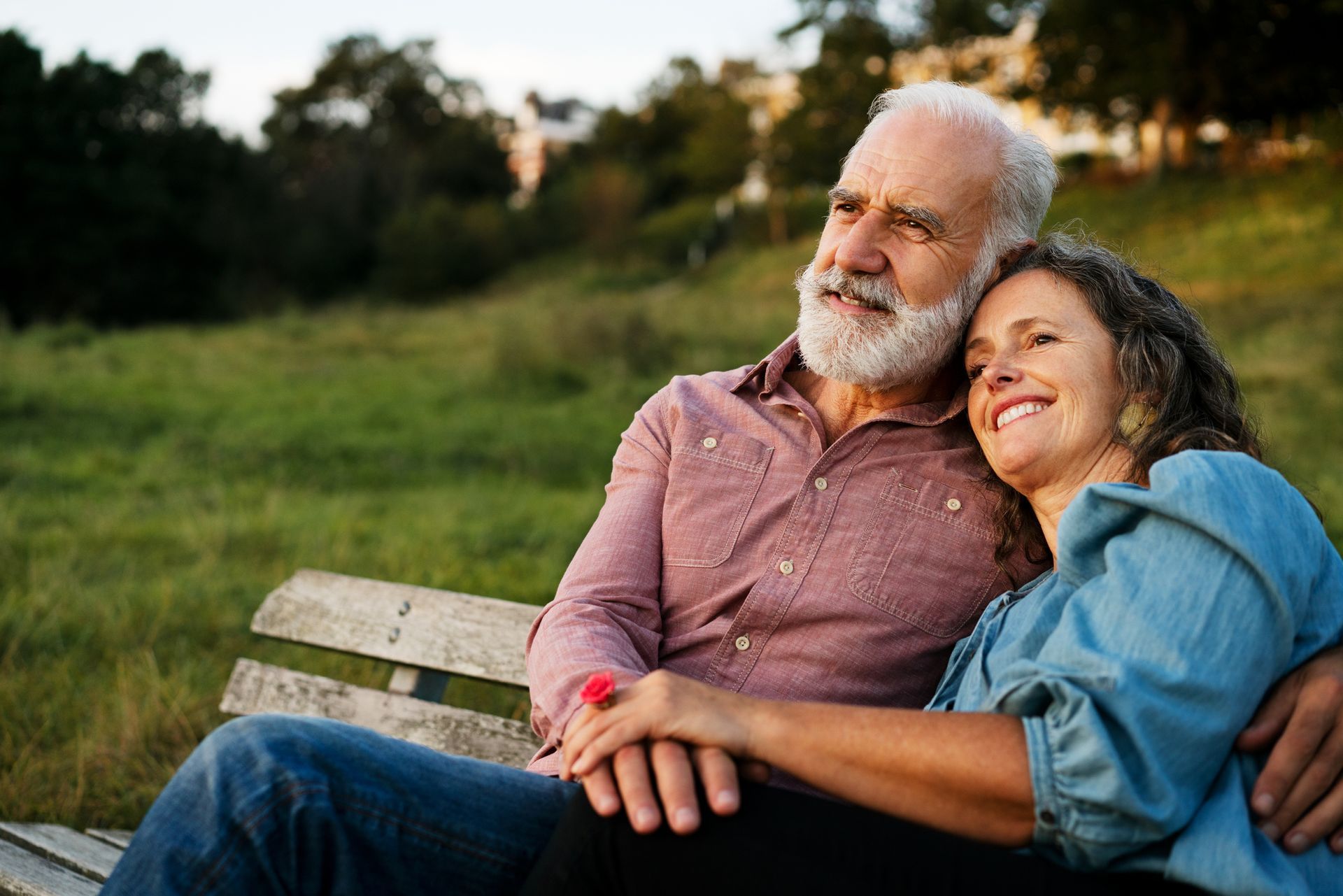 Elderly couple sits on a bench, looking up, with the woman resting on the man's shoulder. Green field, trees in the background.