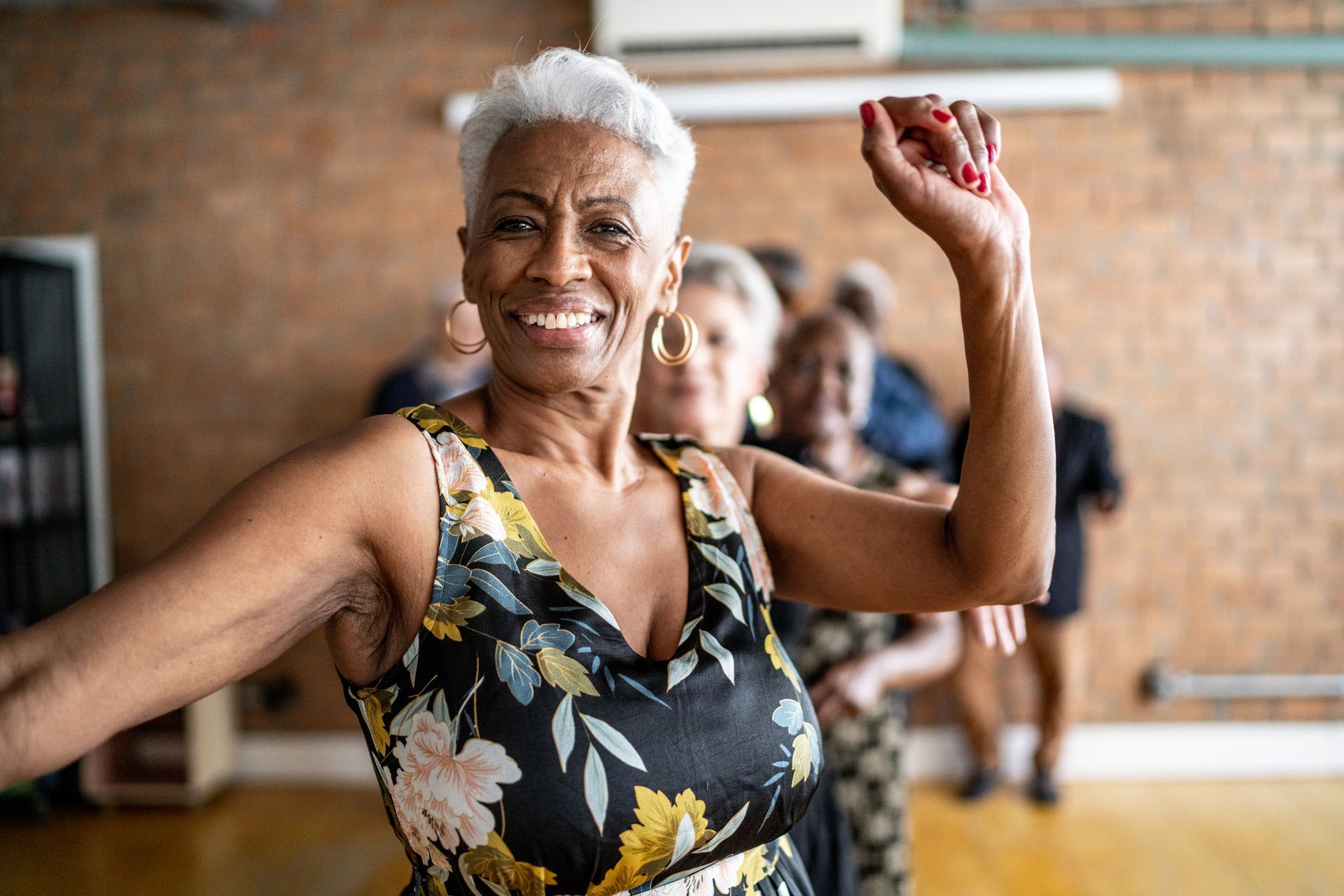 An older Black woman with a gray pixie cut smiles and dances in a dance class.