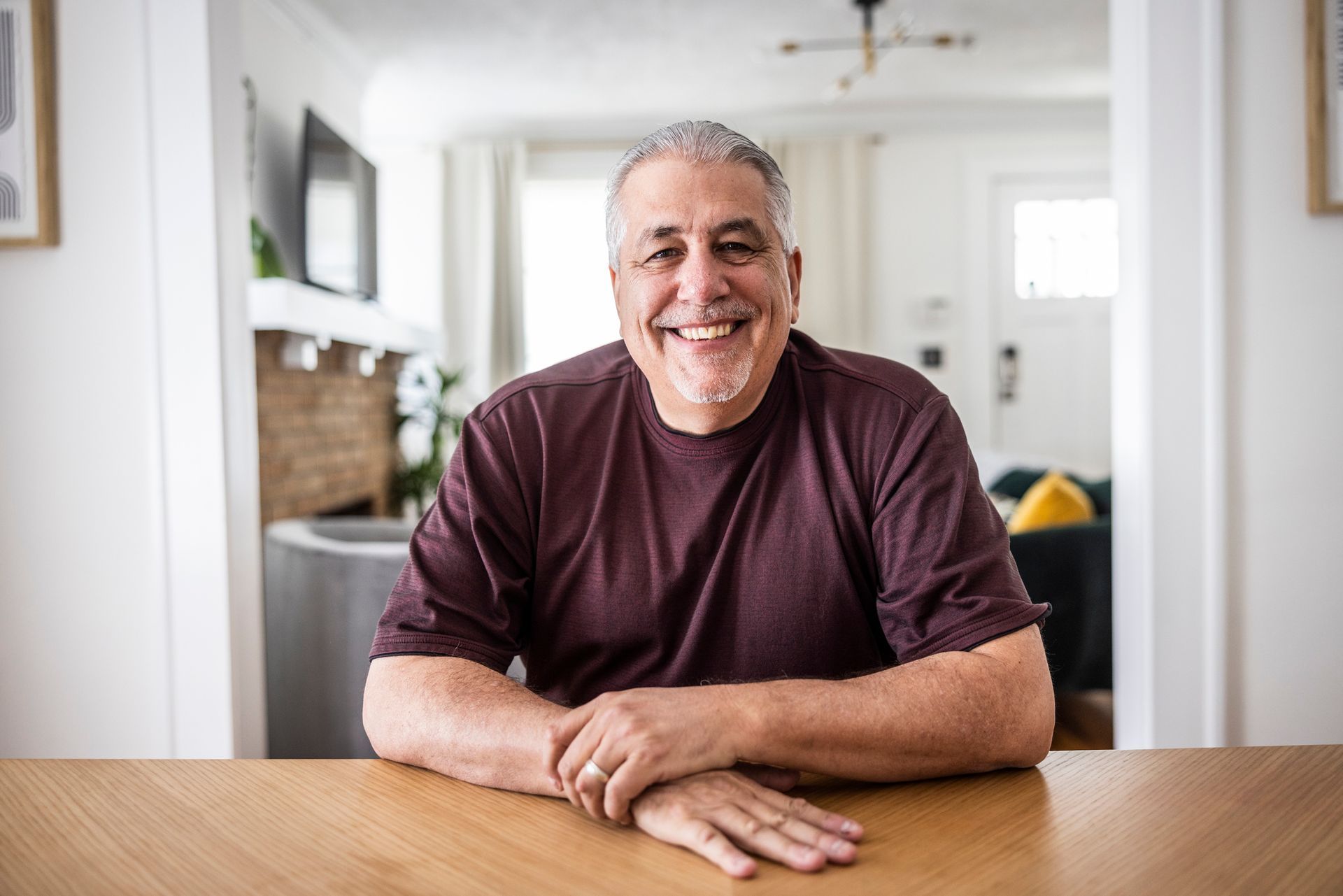 Man with gray hair smiles, sitting at a wooden table indoors.  He wears a maroon shirt.