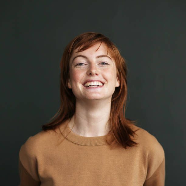 Woman with red hair smiling, wearing a brown sweater, against a dark gray background.