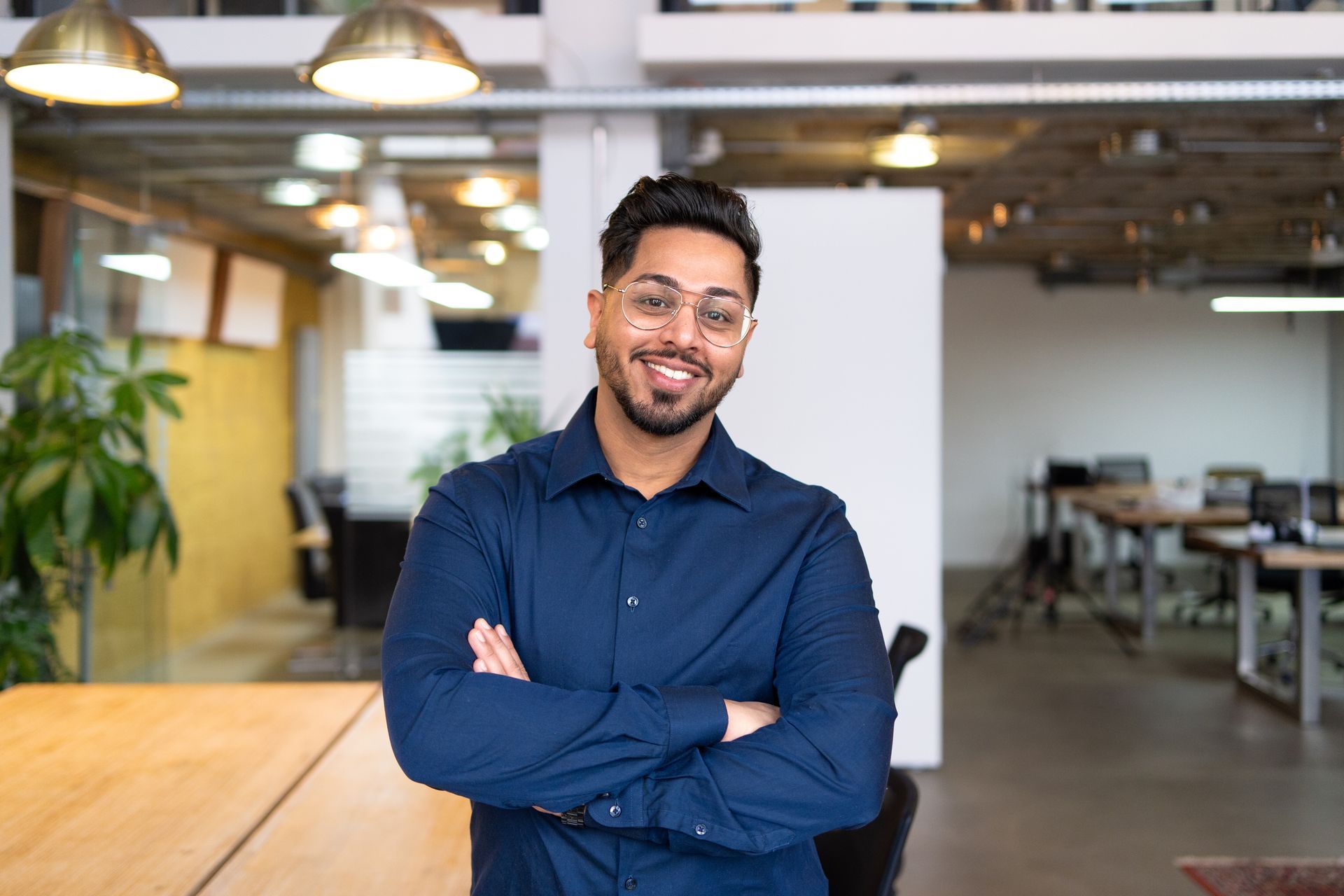 Man in blue shirt smiles with arms crossed in a modern office.