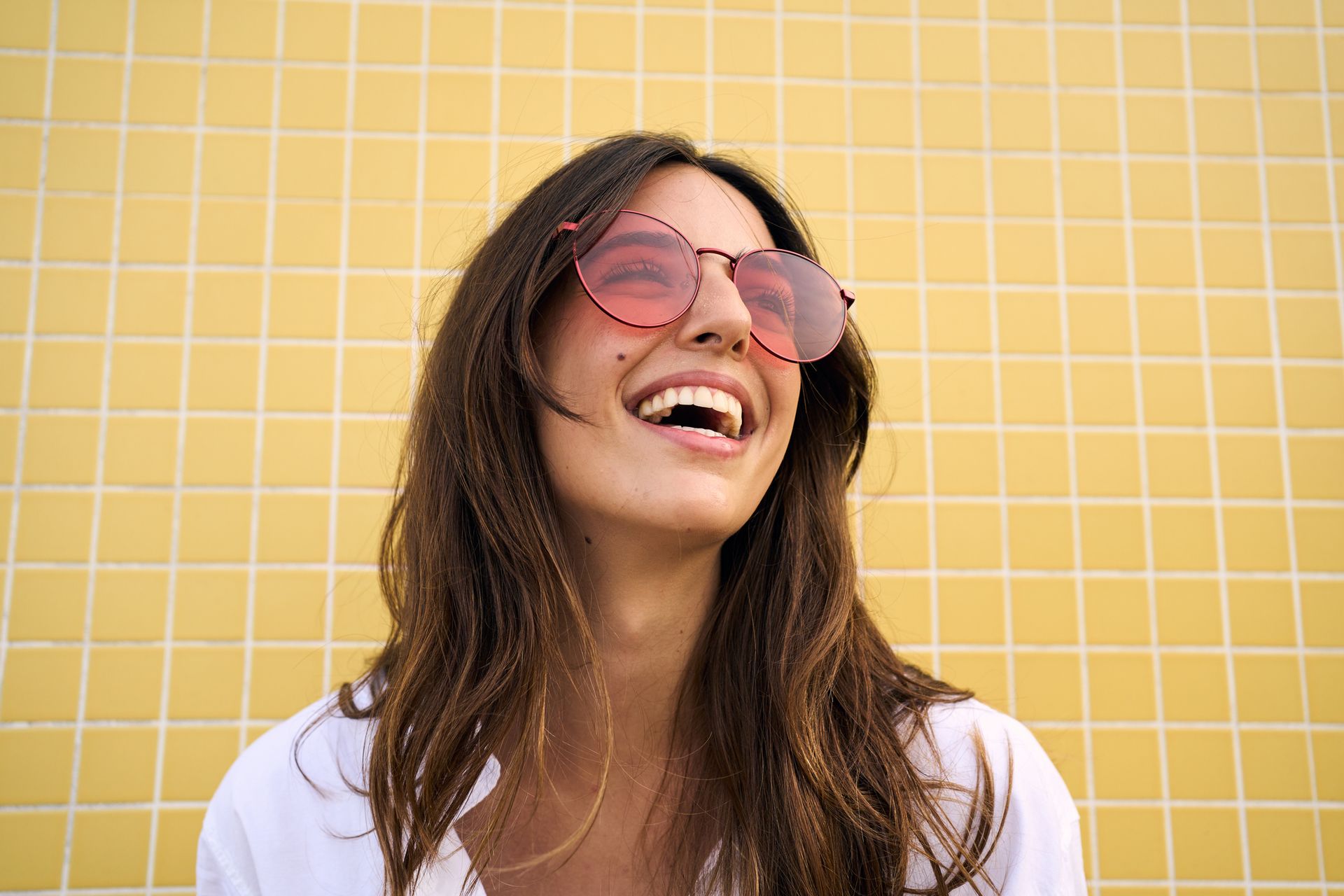 Woman with pink sunglasses laughs in front of a yellow tiled wall.