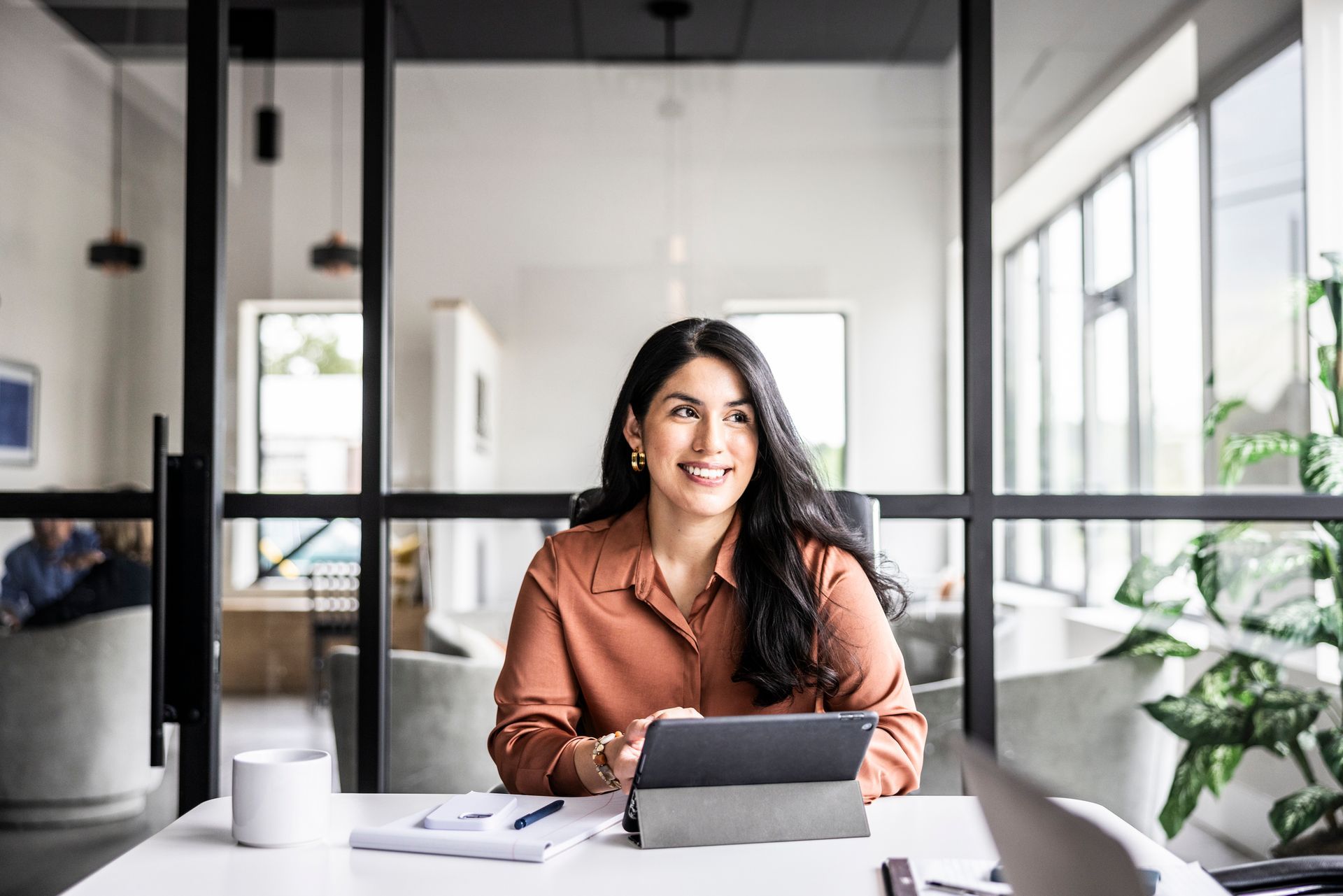 Woman with dark hair smiles, working at a desk in a modern office, holding a tablet.