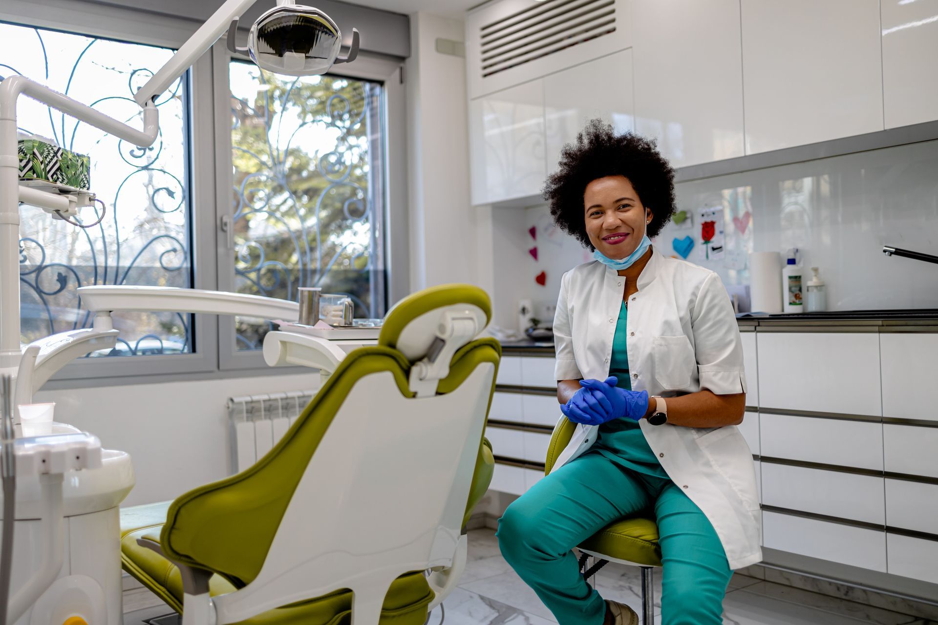 Dentist with curly hair smiles in dental office. She wears scrubs, white coat, and gloves. Green chair, equipment.