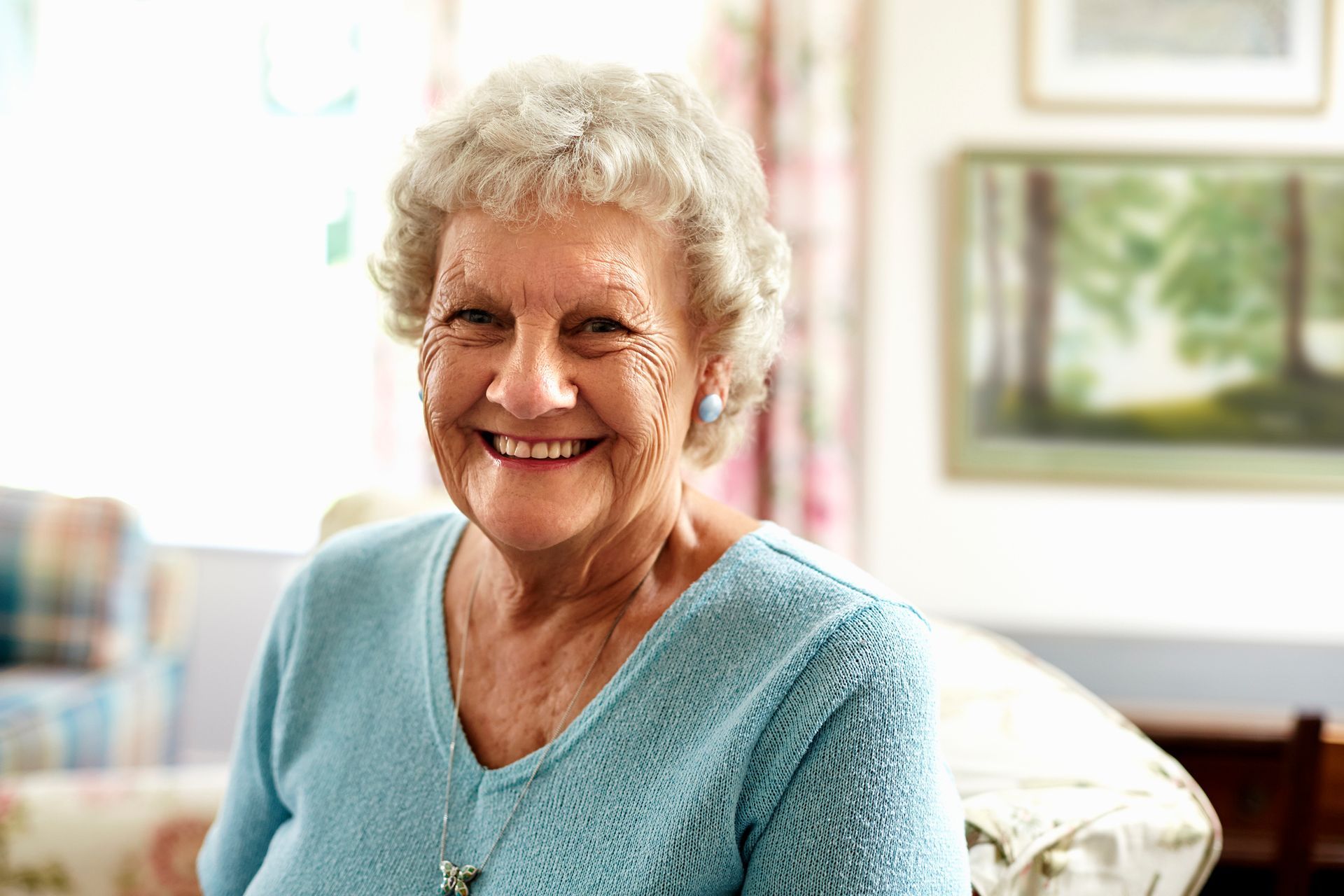 Smiling elderly woman with short gray hair, wearing a blue sweater, indoors near a window and art.