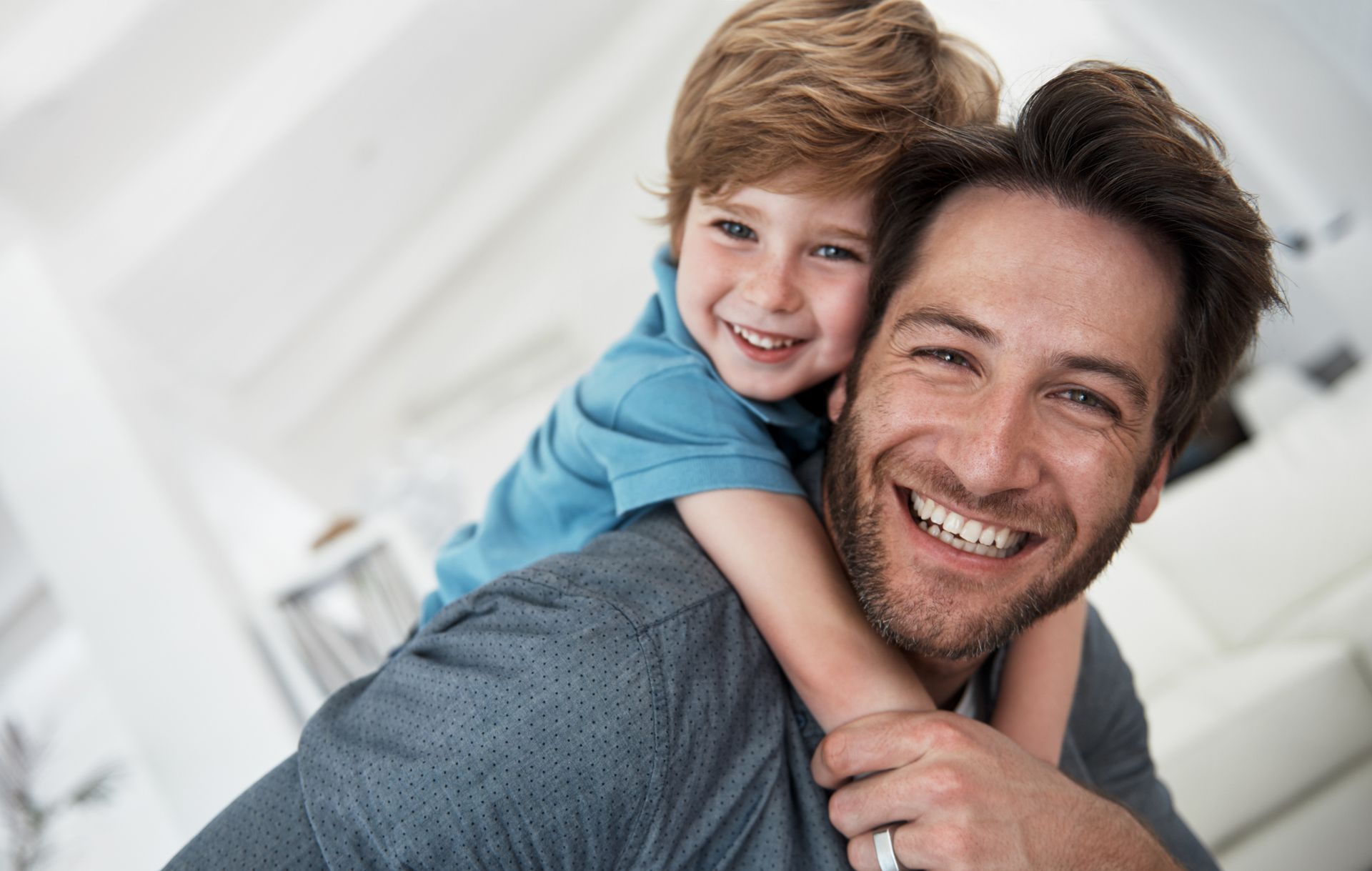 Father giving his smiling son a piggyback ride, inside a bright, airy room.