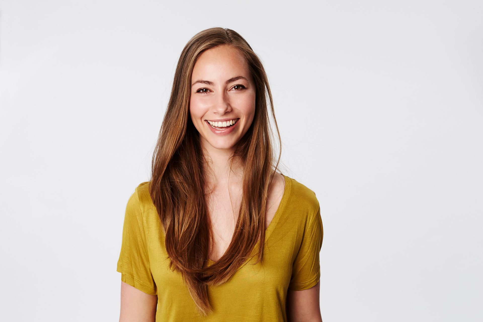Woman with long brown hair, smiling, wearing a mustard yellow shirt, set against a white background.