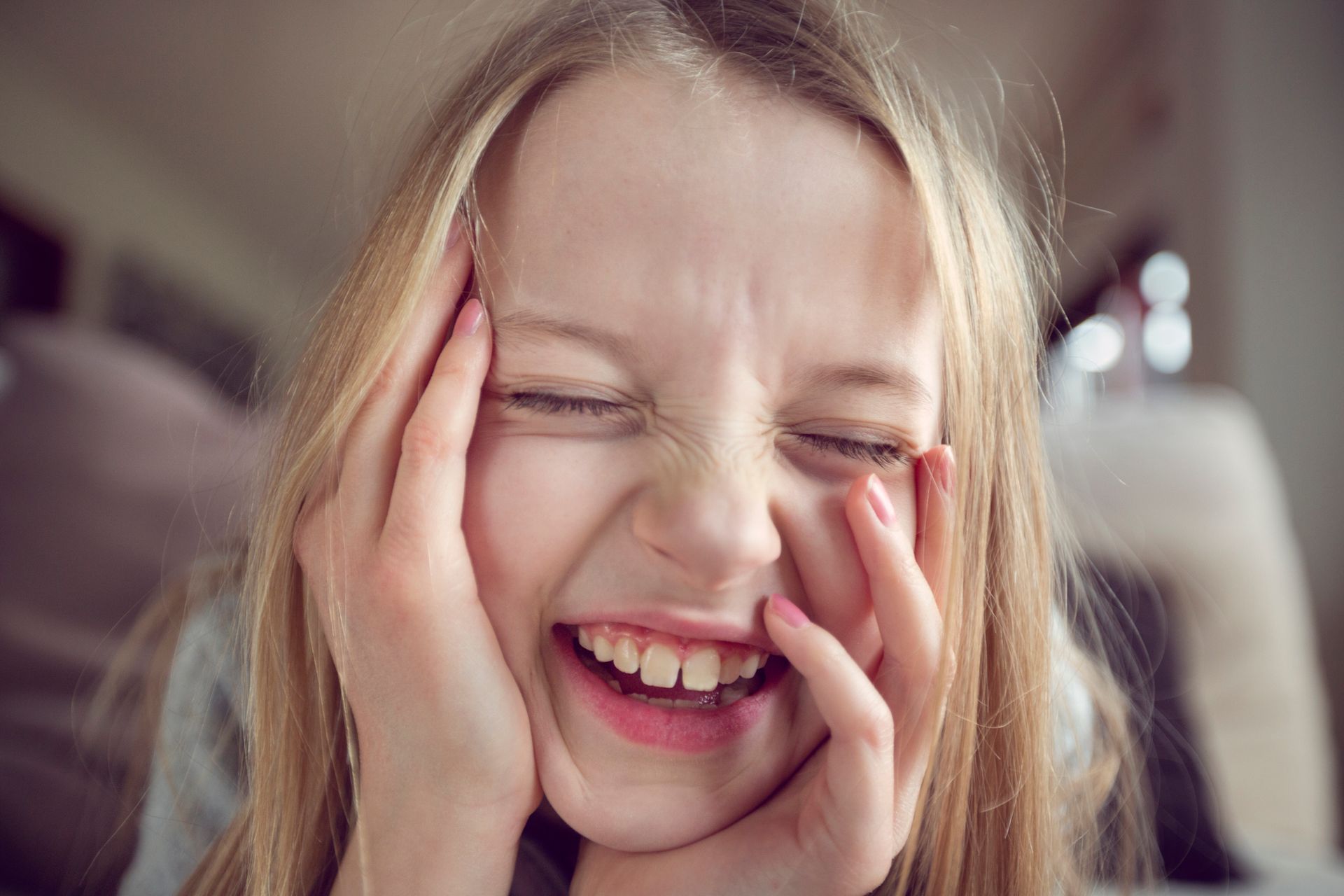 Blonde girl with eyes closed, laughing and holding her face, indoors.