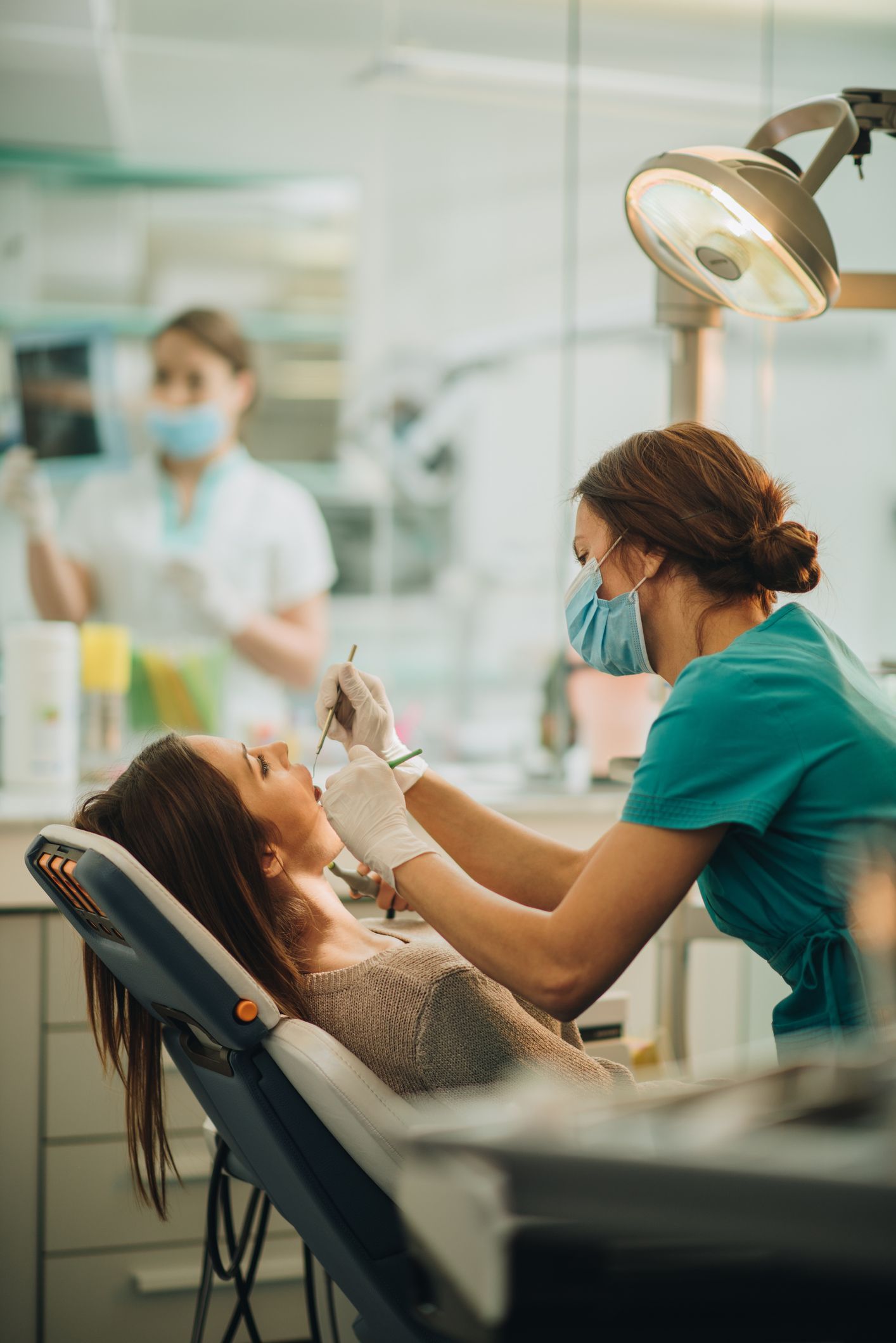 Dentist examining patient's teeth in a dental clinic.  The assistant stands in the background.