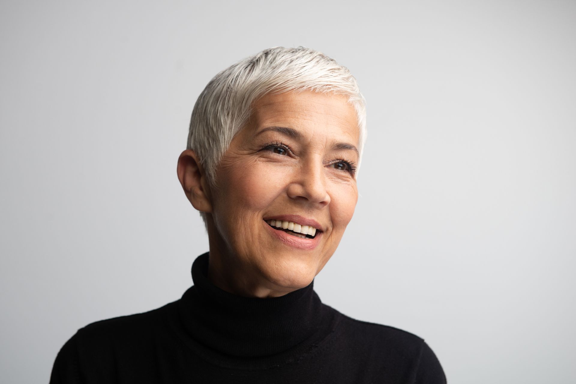 Woman with short gray hair, wearing a black turtleneck, smiles against a gray background.