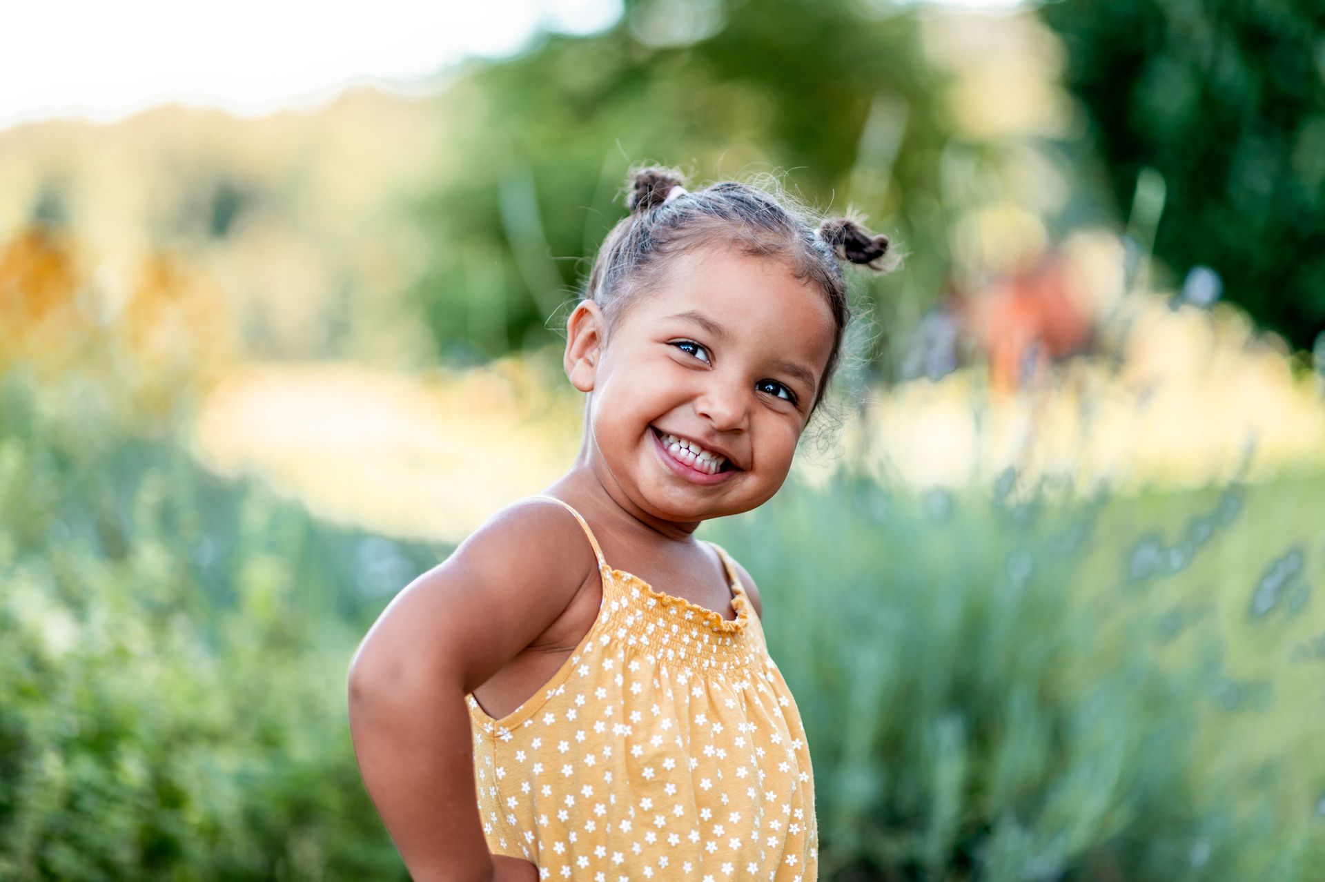 Smiling child with pigtails in a yellow polka-dot dress, hand on hip, outdoors.