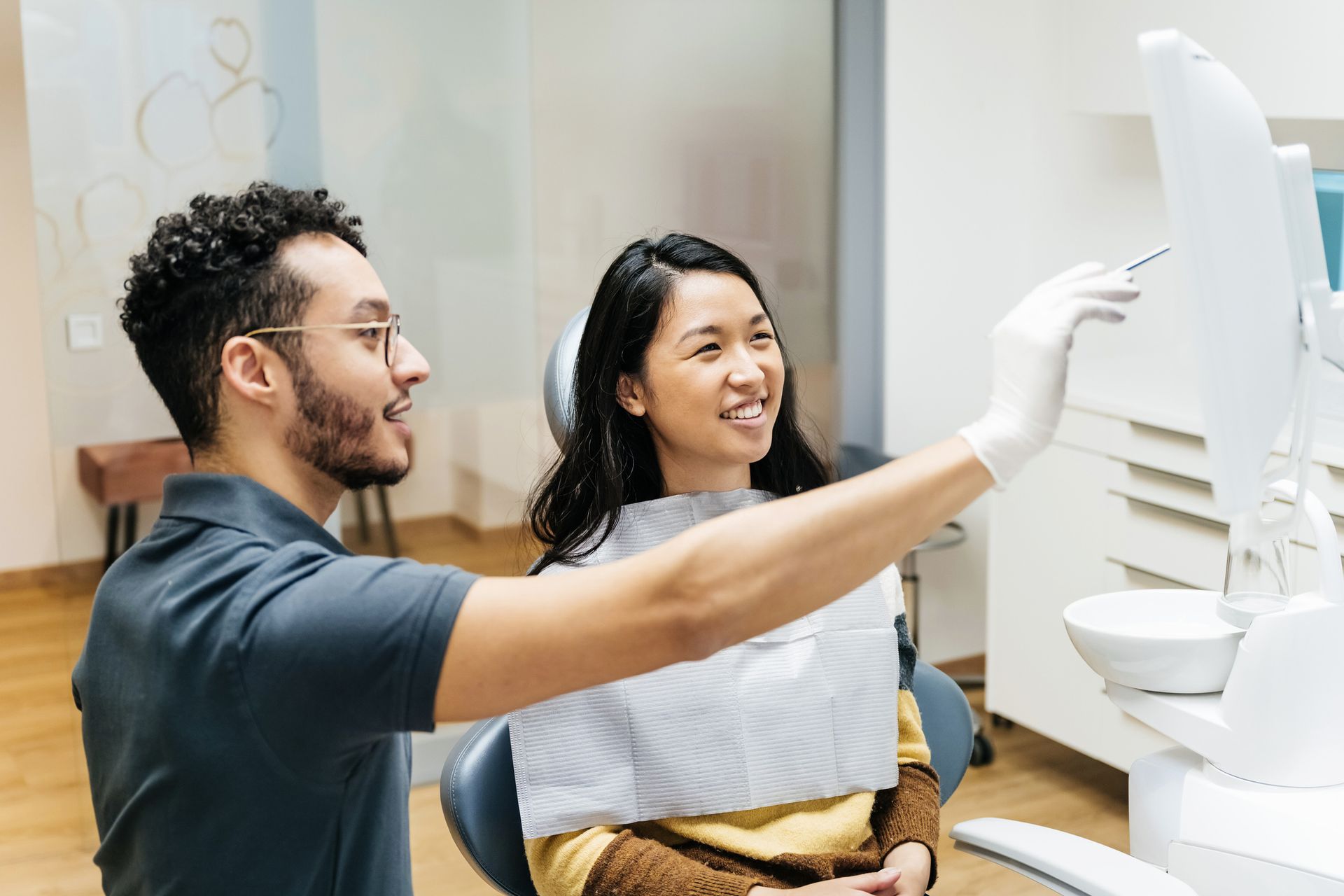 Dentist pointing at a dental mirror, explaining something to a smiling patient in a dental office.