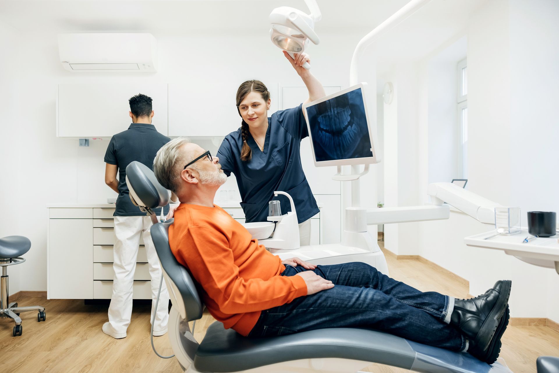 A dentist and assistant with a patient in a dental chair, reviewing an x-ray.