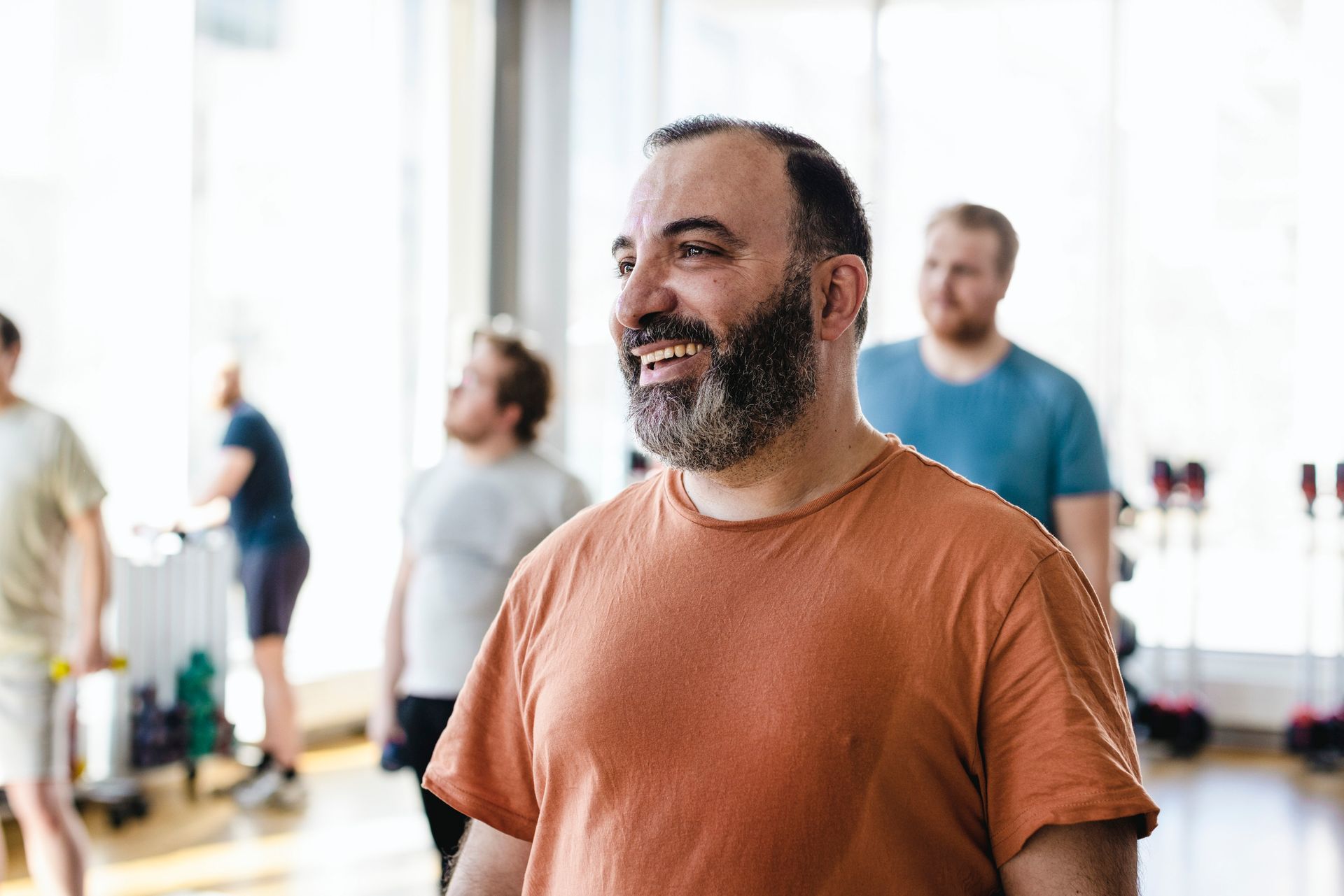 Man with beard smiling, wearing orange shirt, in a gym setting with other men.