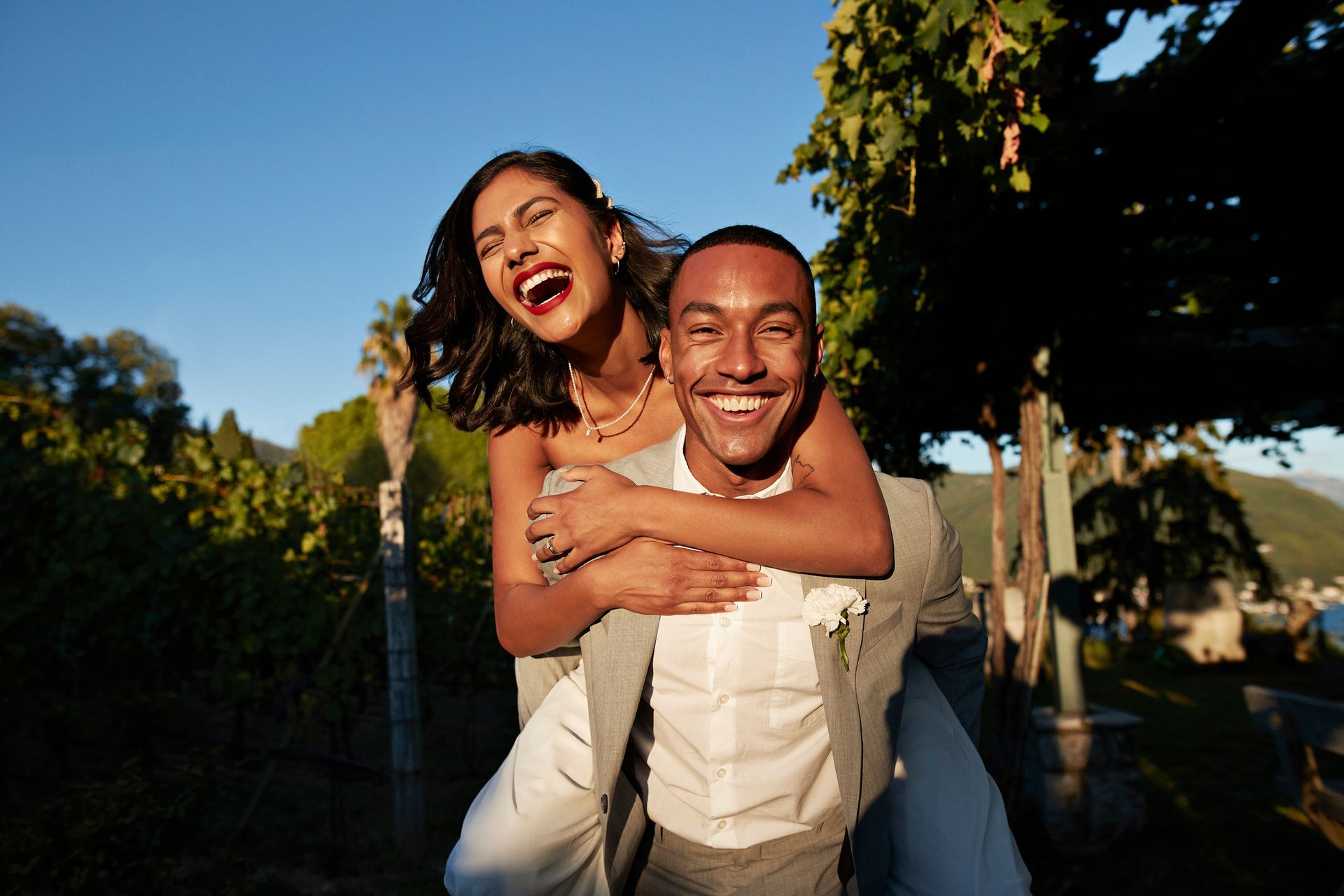 Man giving woman a piggyback ride, both laughing outdoors near a vineyard on a sunny day.