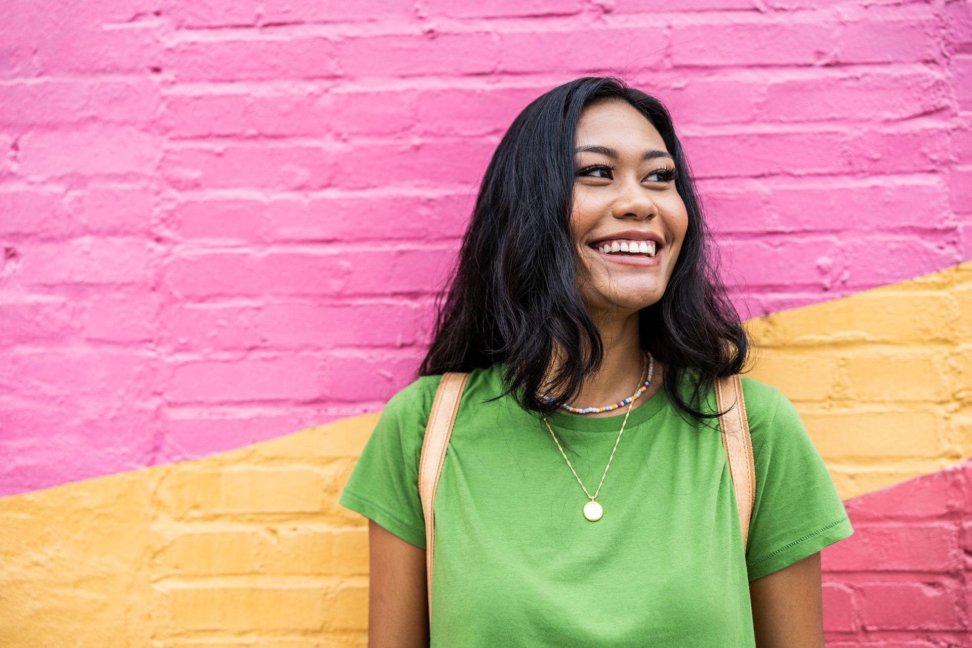 Woman smiling next to a pink and yellow wall, wearing green shirt and necklace.