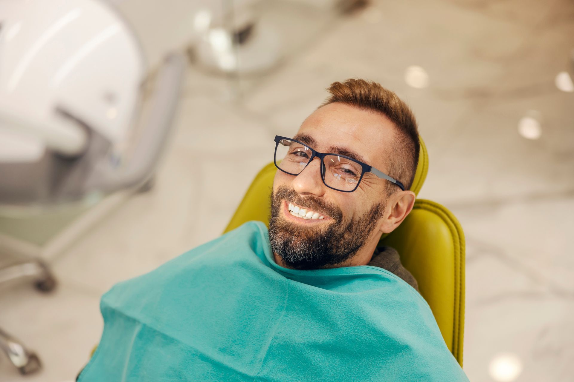 Man in glasses smiles brightly in a dentist's chair, covered by a green bib.