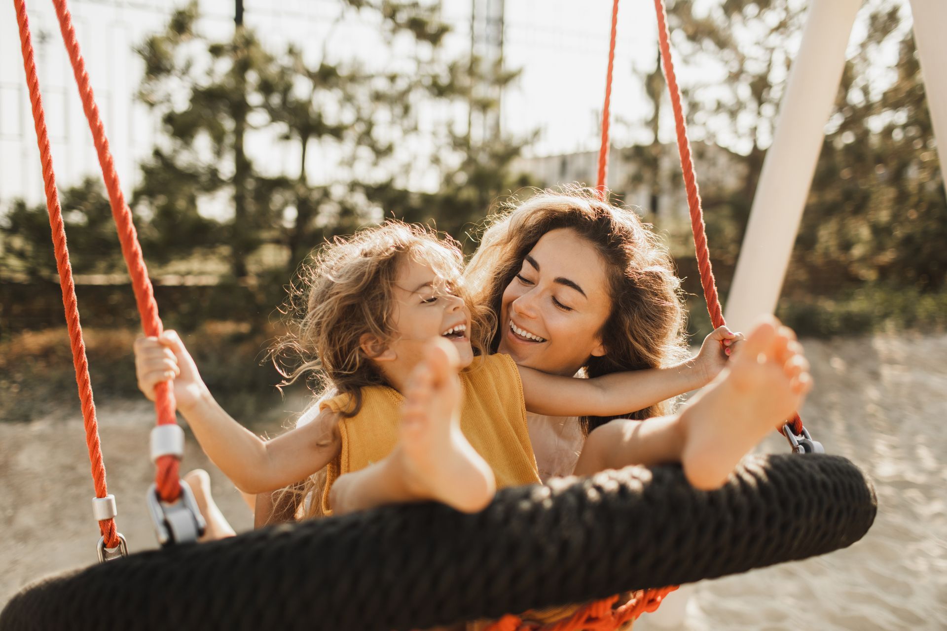 Mother and child laughing on a red swing, outdoors, sunlit, sandy ground.