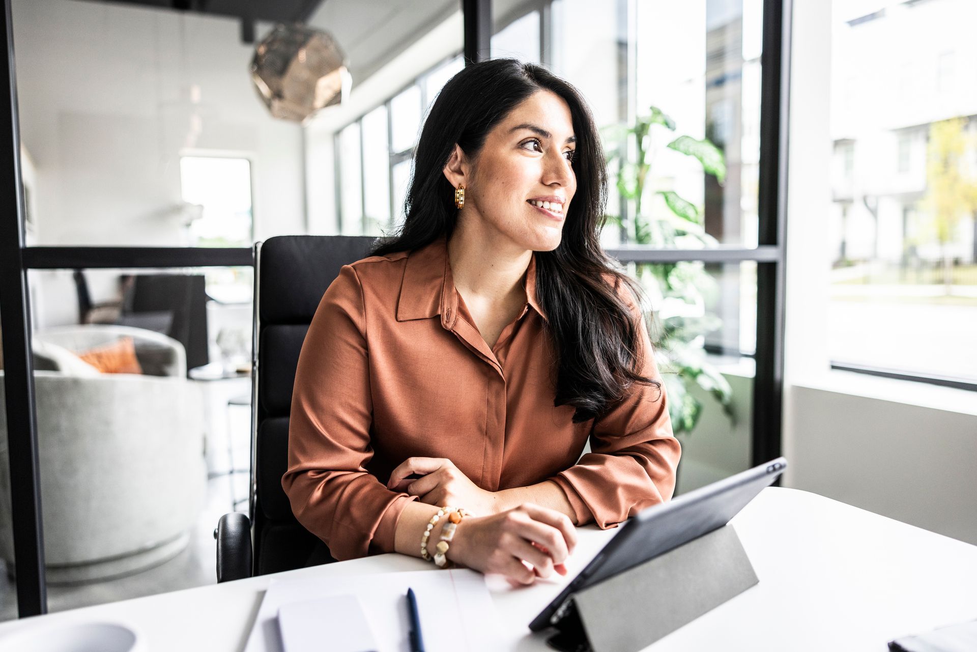 Woman in brown blouse at desk looking out window; modern office setting.
