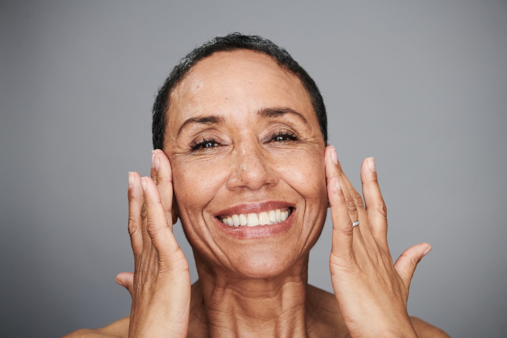 Smiling older woman touching her face, grey background.