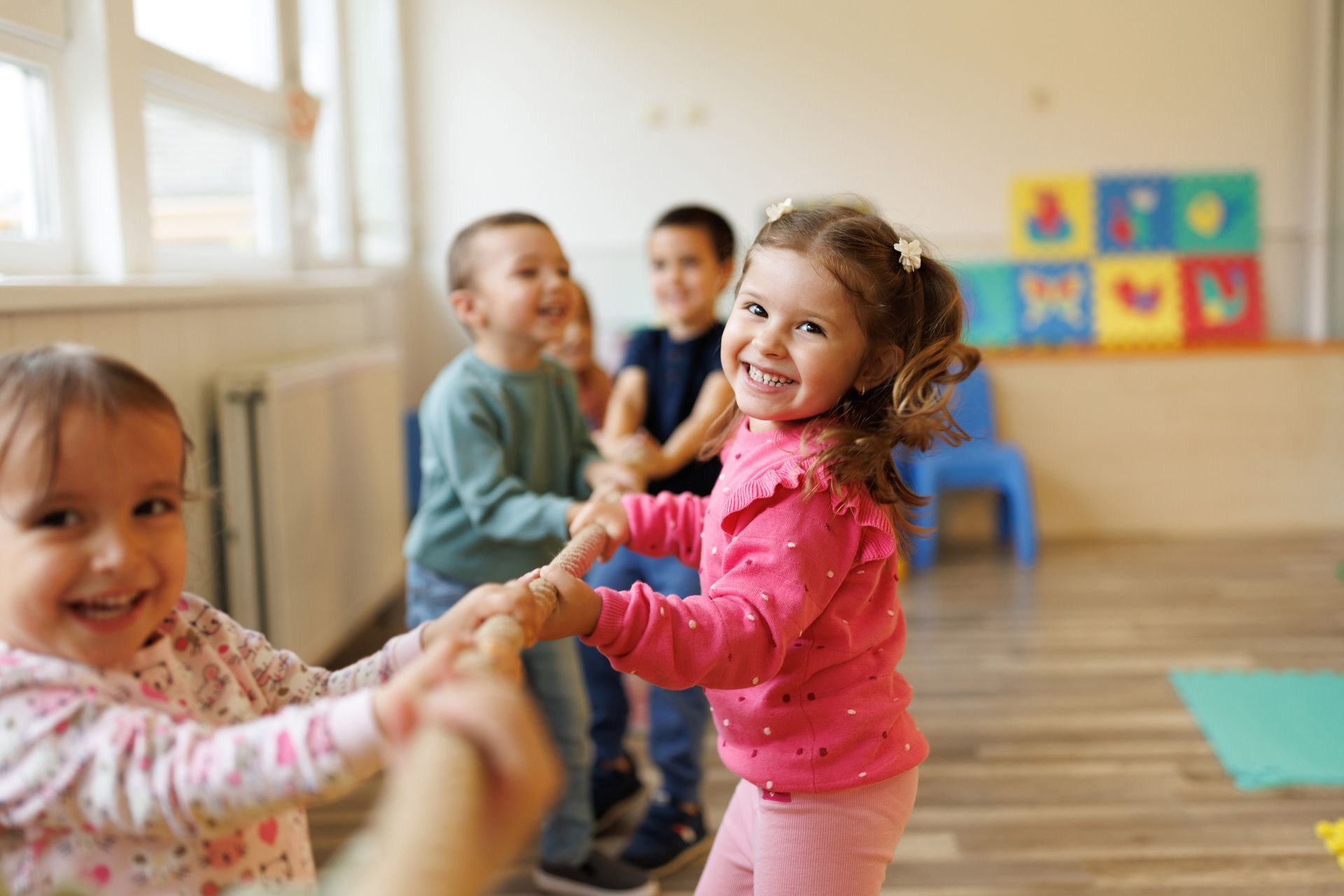 Children laughing and holding hands in a circle, in a brightly lit classroom.