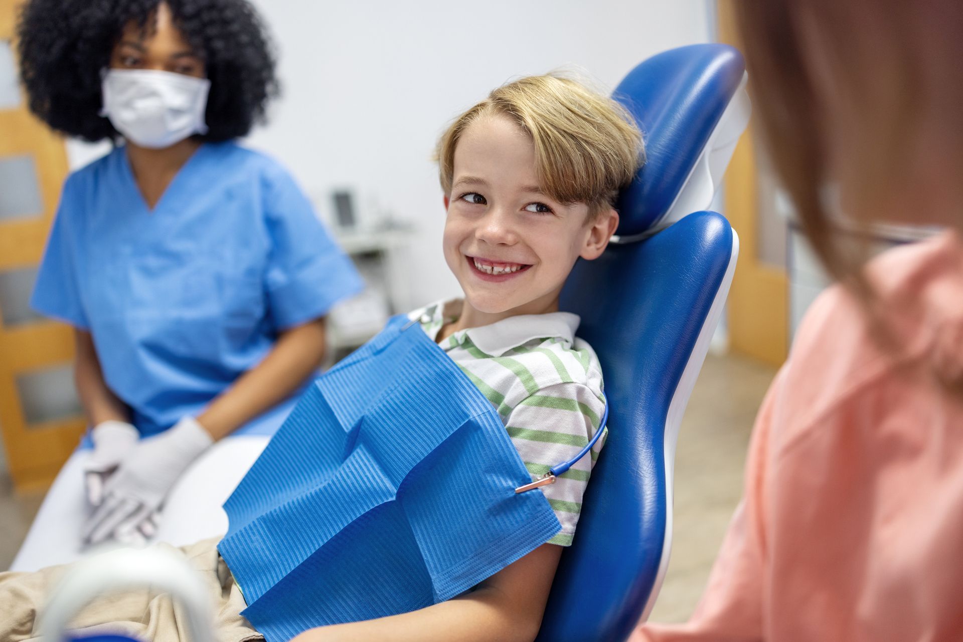 Boy smiles in a dentist's chair; dental assistant and parent present.