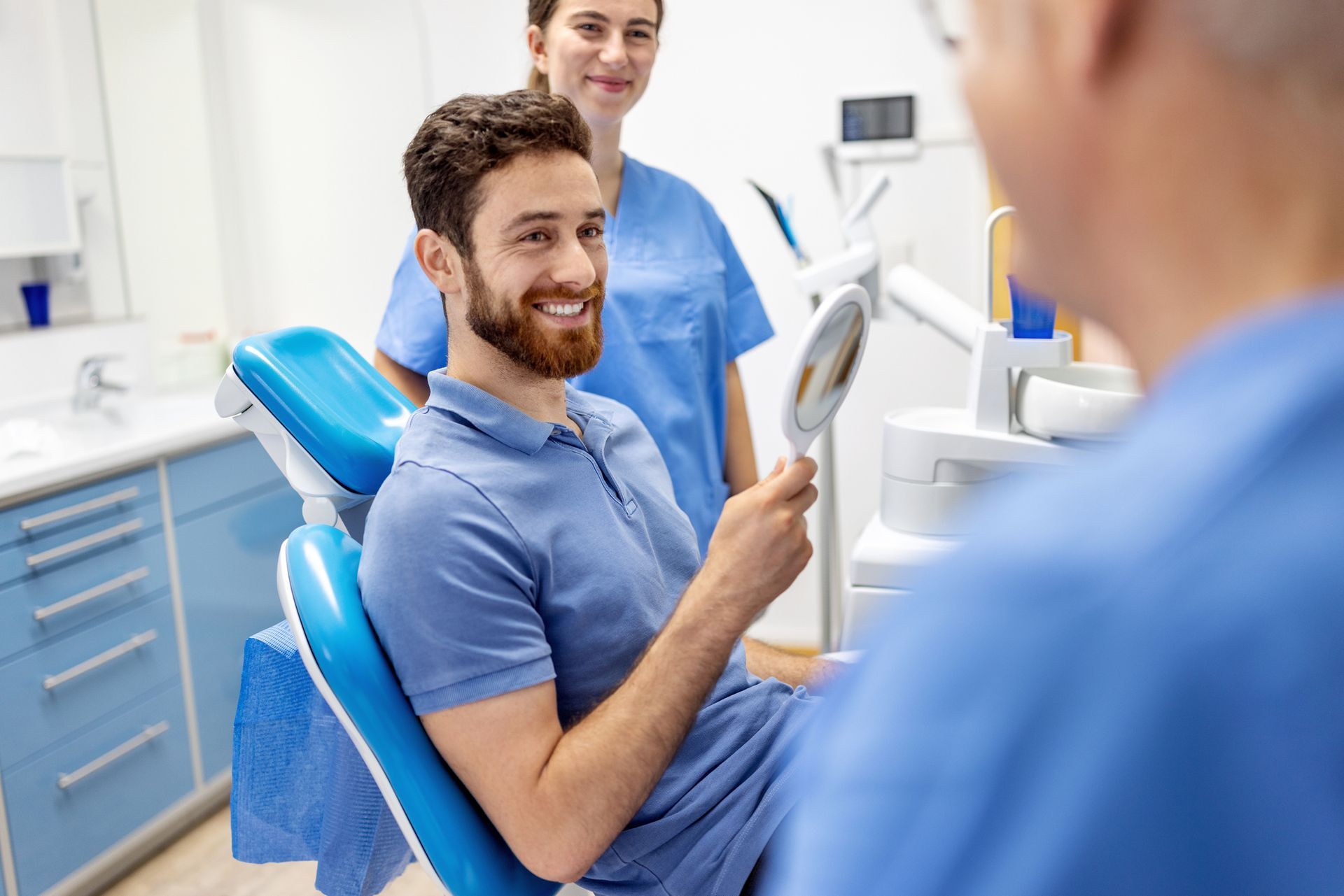 Man in dentist chair smiles, examining teeth in mirror, with dentist and assistant.