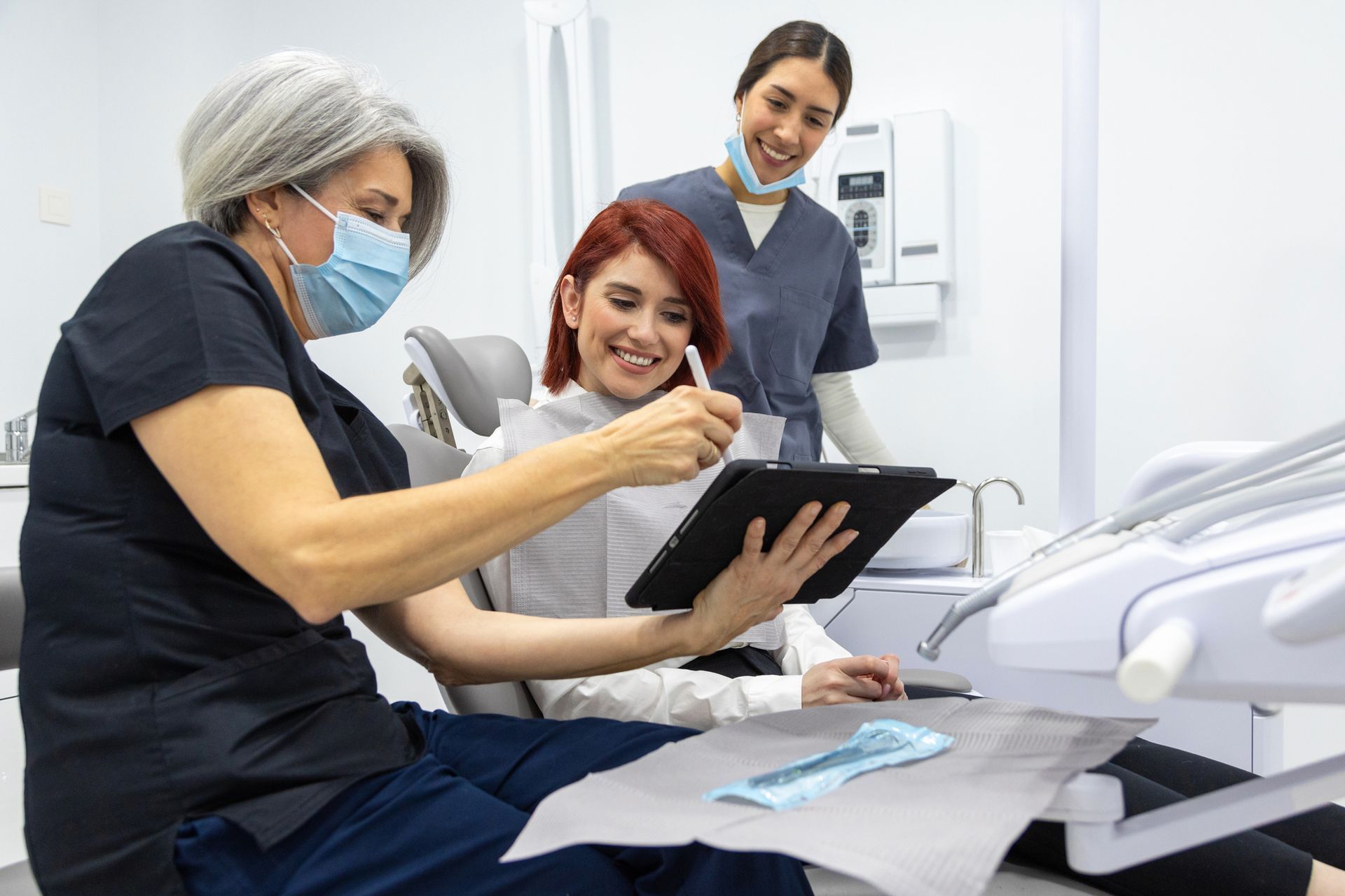 Dentist showing a tablet to a patient, with assistant observing. Modern dental office.